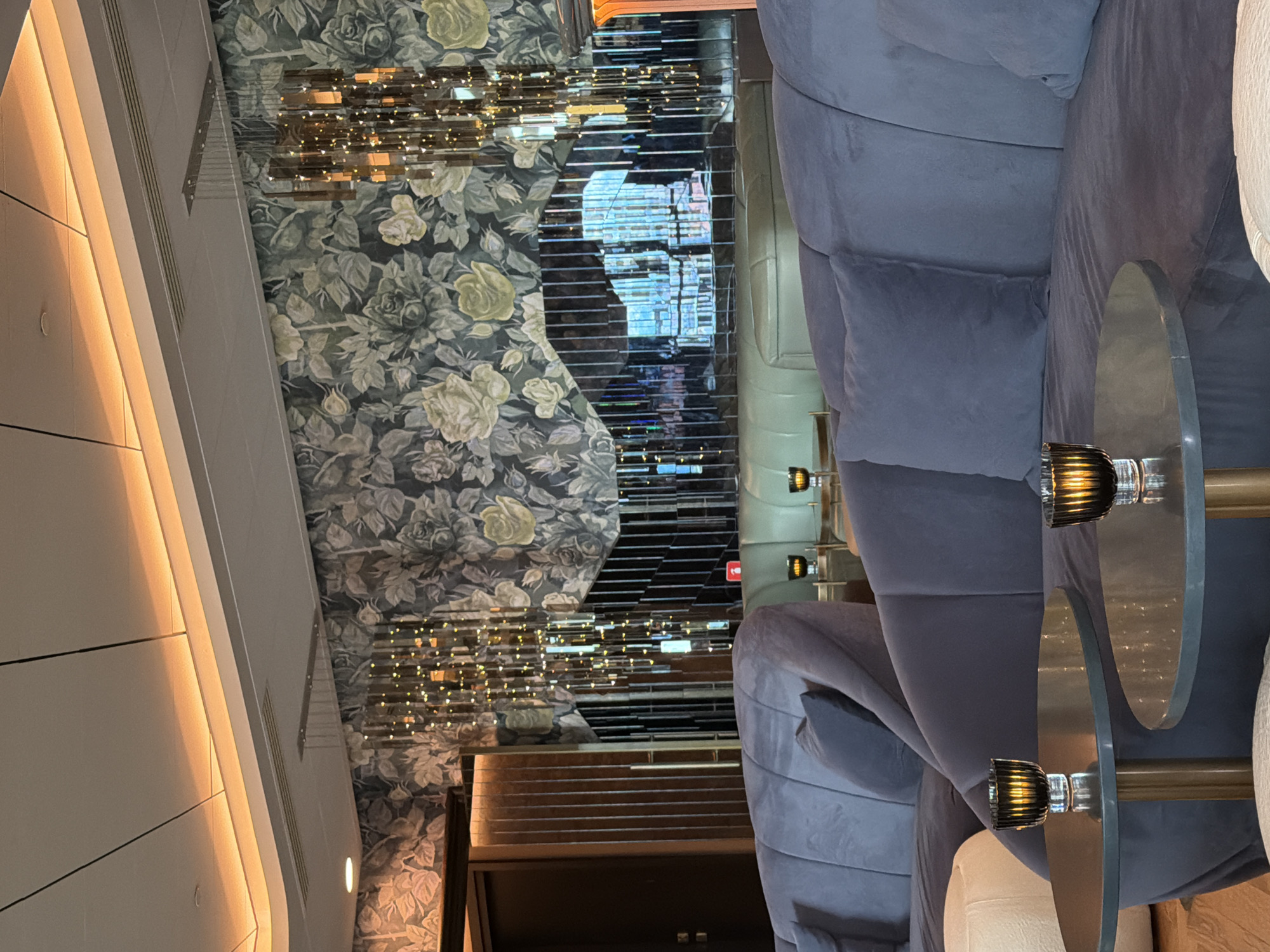 The image shows a dimly lit interior, possibly a bar or lounge in Sofia, Bulgaria. A large, curved, blue velvet couch occupies the right side of the frame, with small, round tables featuring decorative candle holders in front. The wall on the left is decorated with a floral pattern, while the adjacent wall is adorned with a metallic, mirrored, or reflective vertical element. A hallway or doorway is visible in the center. The space is lit by ambient lighting, including what appears to be accent lighting on the wall at left. There are no people visible in the frame.