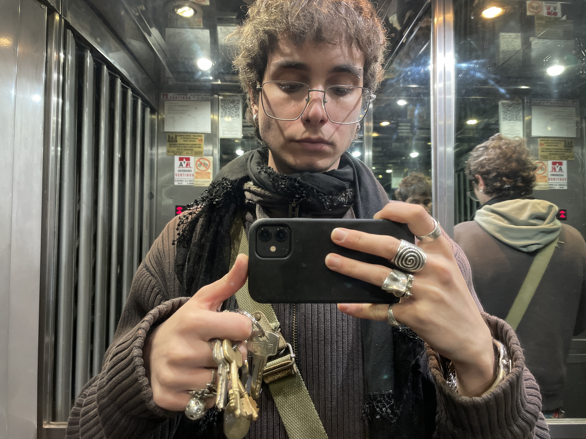 A young person with curly brown hair, wearing glasses and multiple rings, is taking a selfie with a black phone in an elevator. They are wearing a brown sweater, a dark scarf, and a green strap. A bunch of keys are dangling from the hand holding the phone. The background shows the interior of an elevator with ribbed metal walls and mirrored surfaces with several regulatory signs in Spanish, suggesting a location in Buenos Aires, Argentina. The reflection in the mirror shows the person's back and the details of the elevator interior.