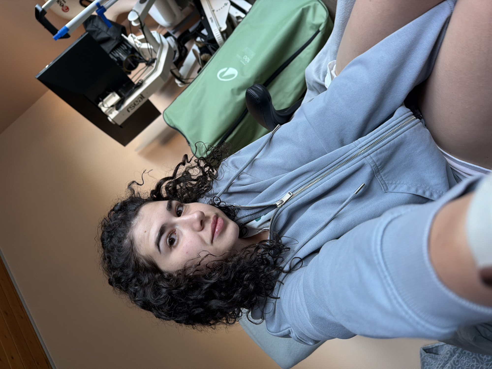 A young woman with curly dark hair is sitting in a chair, looking towards the camera. She is wearing a light blue zip-up hoodie. Behind her, a green bag and medical equipment are visible. The background appears to be an indoor setting with a wall and ceiling made of wood panels. The lighting is warm and diffused. There is no indication of the location in Sofia, Bulgaria.