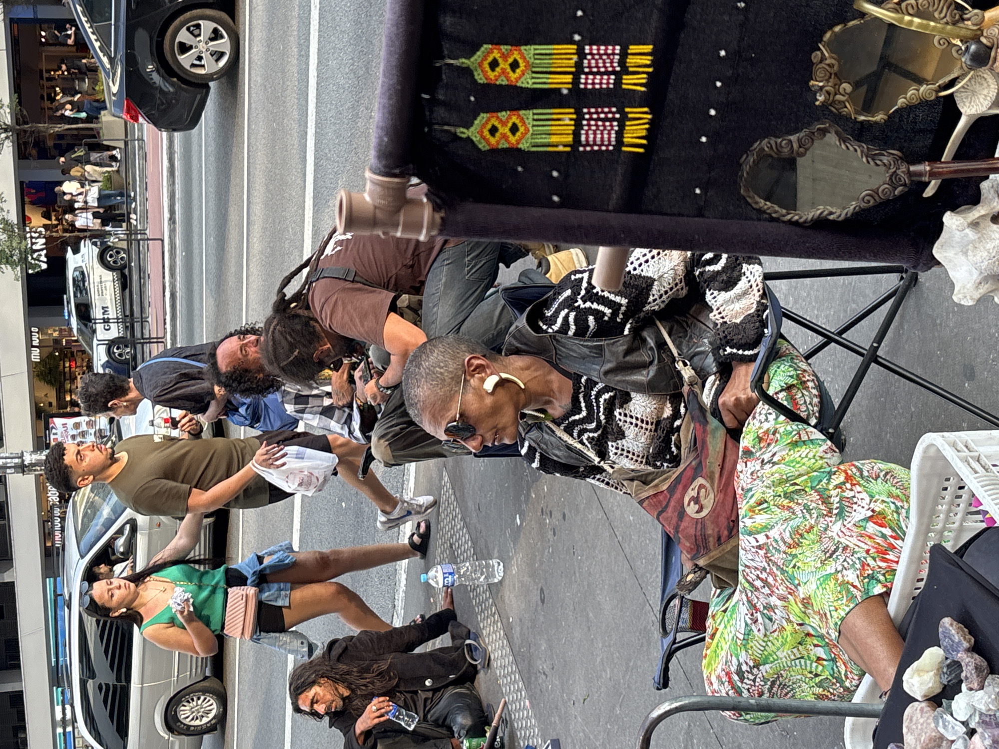 The image shows a street scene in São Paulo, Brazil, with a pedestrian sidewalk lined with a street vendor's display of handcrafted items and mirrors, and various people passing by. A vendor sits on a folding chair next to a table draped in a black cloth with yellow and red designs. On the table are small mirrors and bone or shell-like objects. The vendor is wearing sunglasses and a patterned jacket. Customers are standing around the vendor. A street vendor sits on the ground, a water bottle next to him. Pedestrians walk behind them, next to a row of parked vehicles. Buildings are visible in the background.