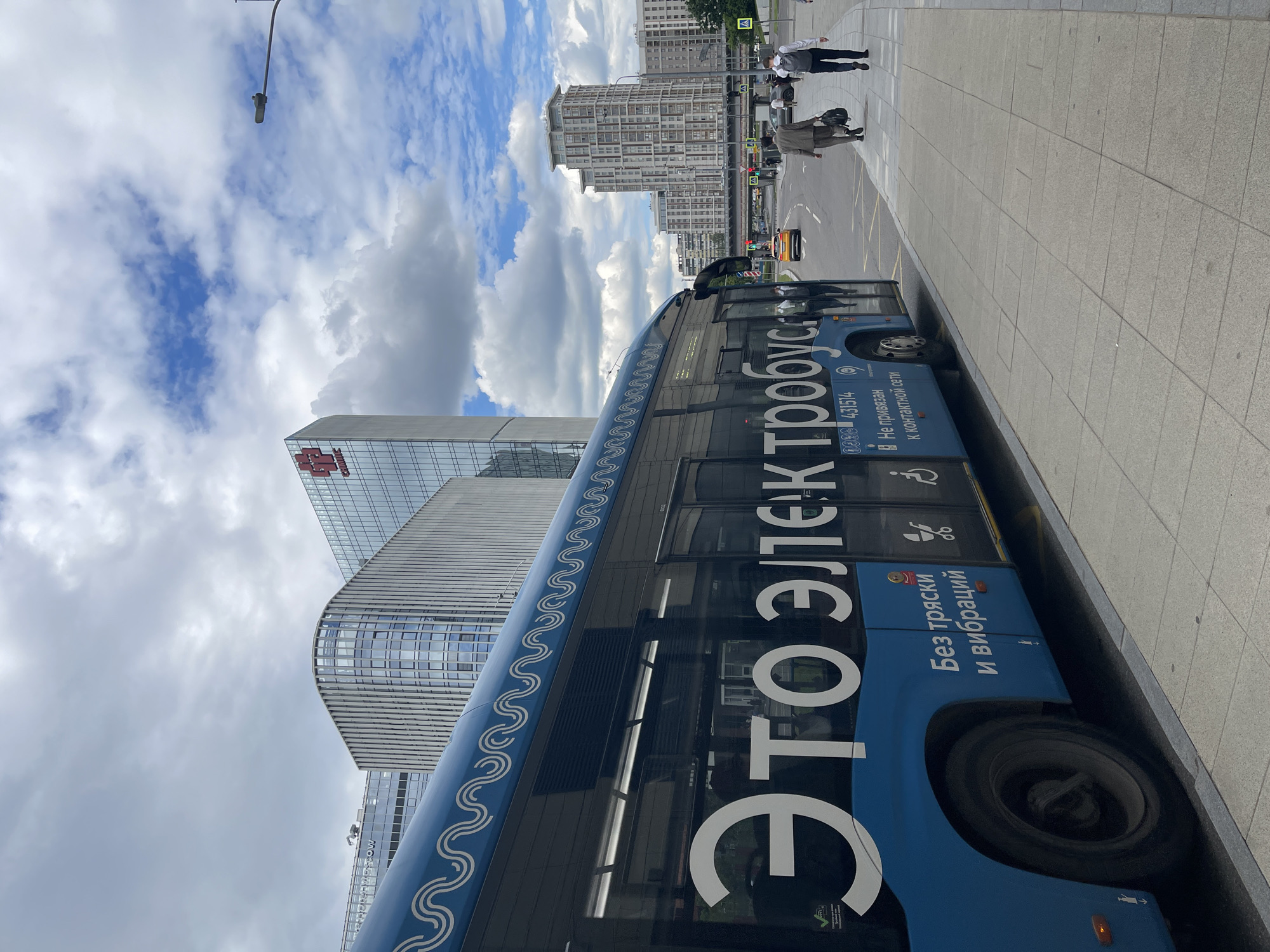 A blue electric bus is parked on the side of a street in an urban environment. The bus has white text on its side that reads "ЭТО ЭЛЕКТРОБУС". Below this text, smaller text reads "Без тряски и вибраций" and "Не привязан к контактной сети". There are also icons for a stroller and a person in a wheelchair. The bus is partially obscuring the view of several modern buildings in the background. A sidewalk with grey paving stones is in the foreground, and a few people are walking on it. A yellow car is driving down the street. The sky is partly cloudy.