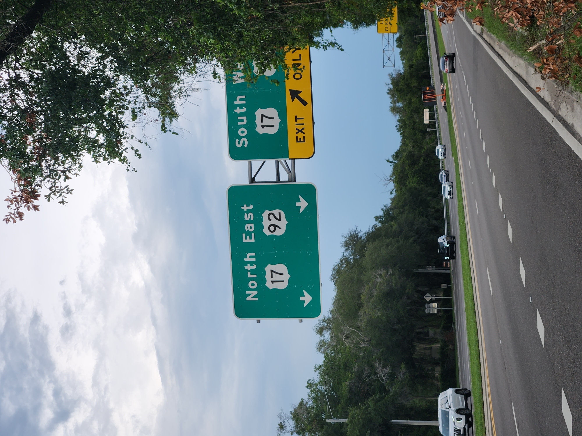 The image shows a highway scene in Maitland, Florida. The perspective is from a moving vehicle, looking towards the right. Overhead signs indicate directions. The top sign, partially obscured by trees, directs traffic to "South W", "17", and "EXIT ONLY" with an arrow pointing right. Below it, another sign reads "North East", "17", "92", and has an arrow pointing right. Several cars are visible on the multi-lane highway, some in the distance and one in the foreground. The highway is flanked by a grassy shoulder and dense trees on the left and right. The sky is partially cloudy, suggesting it is daytime, likely morning or afternoon. The overall impression is of a typical highway commute on a clear day in a suburban or rural area of Florida.