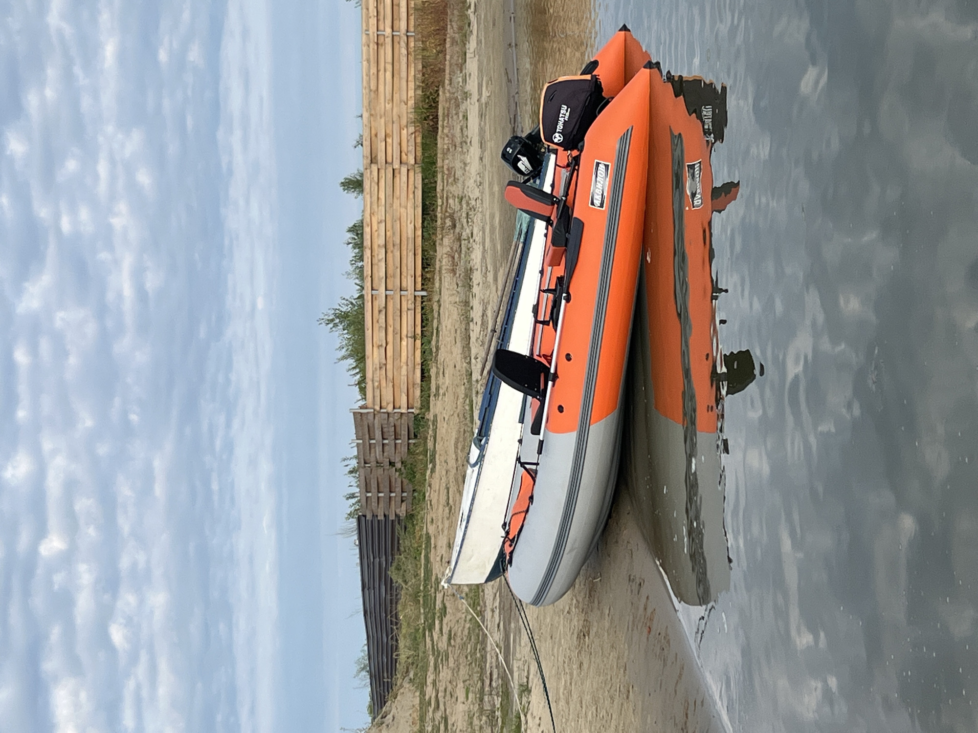 Two inflatable boats are beached on a sandy shore beside a body of water. The foremost boat is orange and gray, with an outboard motor at the stern. The second boat, white with blue trim, is positioned behind and slightly to the left of the orange boat. A wooden fence runs along the top of a sandy embankment behind the boats, with some sparse vegetation visible. The sky is overcast with a pattern of gray clouds. The water reflects the boats and the sky. The location is identified as Novo-Pichyugo, Russia. There are no people visible in the image.