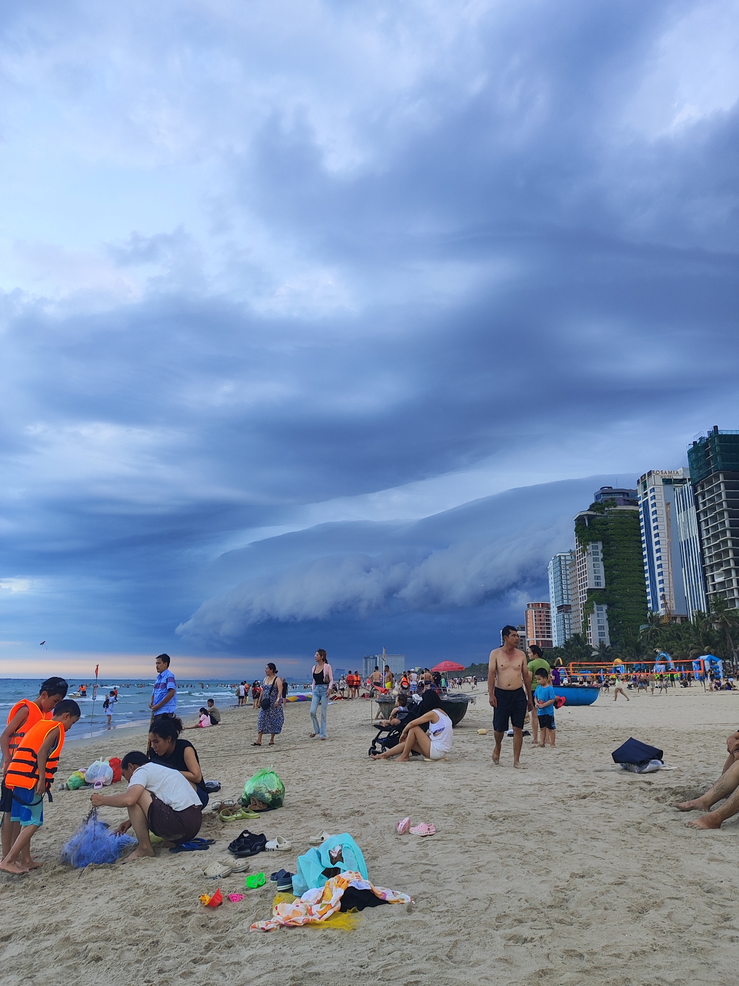 This is a wide-angle, eye-level shot of a crowded beach in Da Nang, Vietnam. The sky is filled with dramatic, dark storm clouds, with a distinct shelf cloud formation visible over the ocean. In the foreground, people are scattered across the sandy beach. Several children in orange life vests are interacting with a blue fishing net. Adults are seen sitting, standing, and walking. In the background, high-rise buildings line the coast, some adorned with greenery. The ocean is calm, with some boats visible further out. The overall atmosphere suggests an approaching storm.