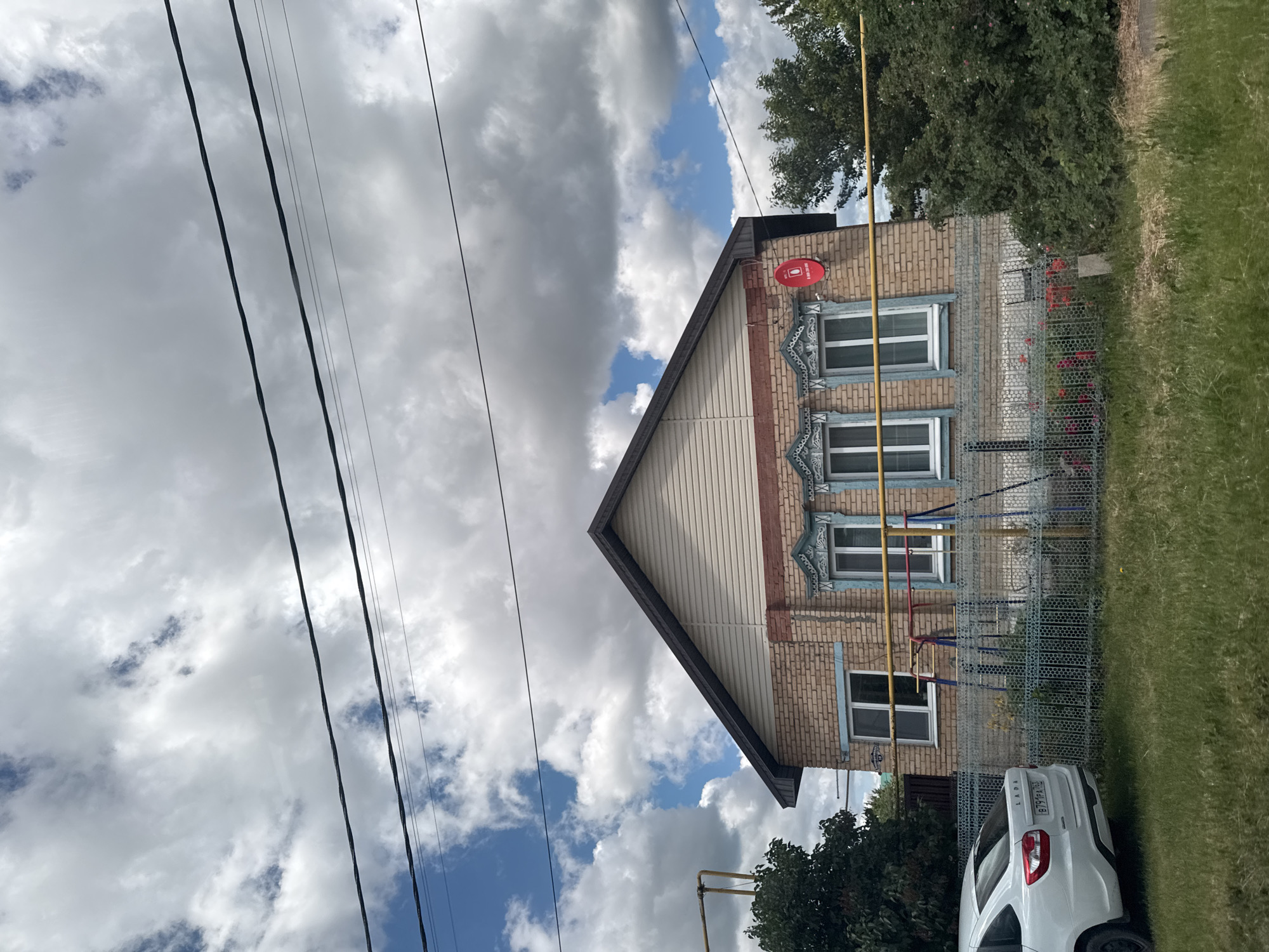 The scene depicts a two-story house in an unidentified urban location. The house is constructed of tan brick with wood siding on the upper gable end. Three windows with decorative, light-blue trim are visible on the brick section of the house. A red satellite dish is mounted on the upper corner of the house, near the roofline. A chain-link fence runs along the side of the house, with green grass growing behind it. A white car is parked partially in the frame, near the fence. Power lines are visible in the foreground, stretching diagonally across the image. The sky is filled with white and gray clouds, with patches of blue visible between them.