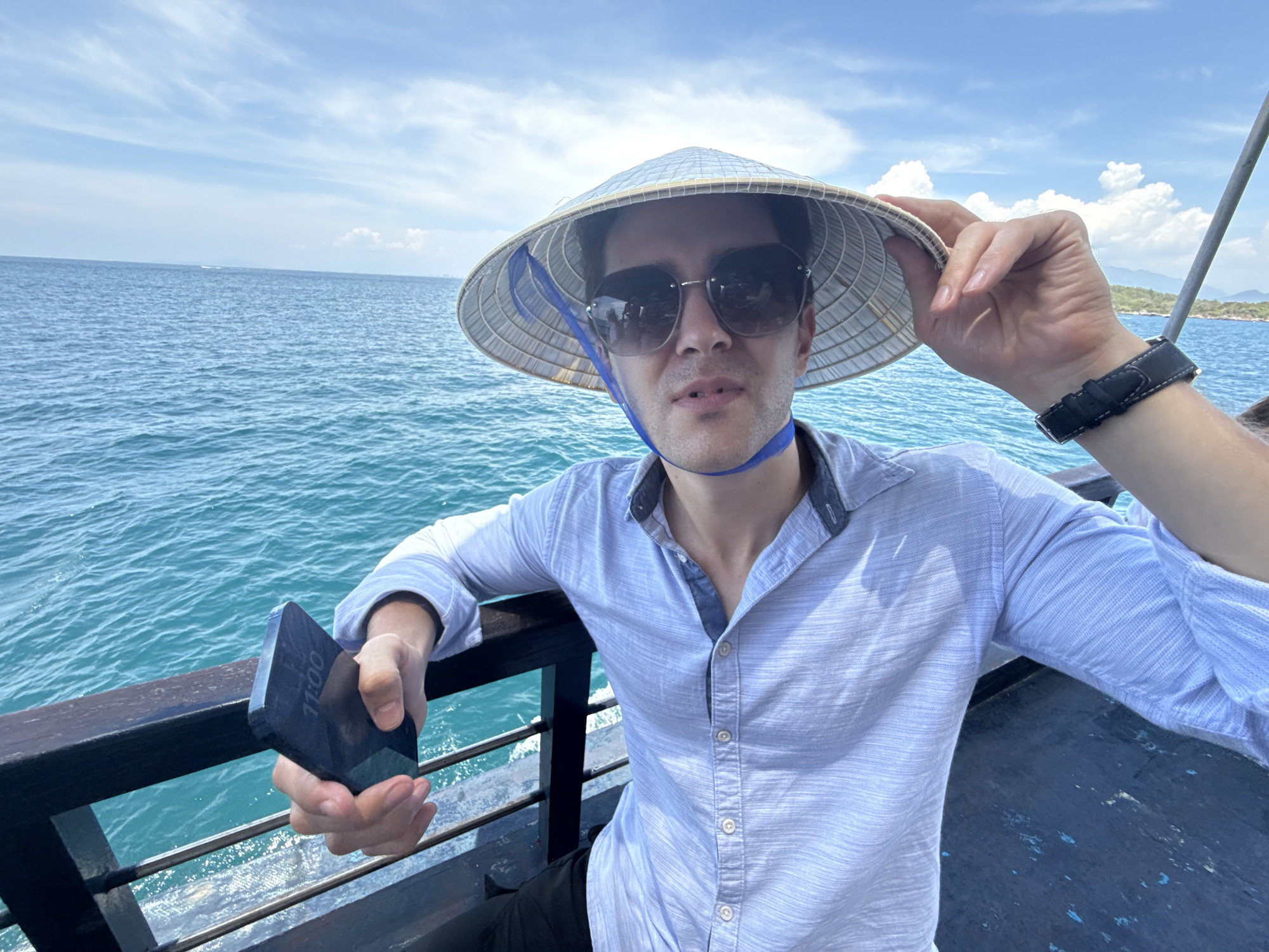 A man is on a boat in Nha Trang, Vietnam, wearing a traditional conical hat and sunglasses. He is holding a mobile phone, and grasping the brim of the hat with his other hand. The ocean, sky, and distant coastline are visible in the background.