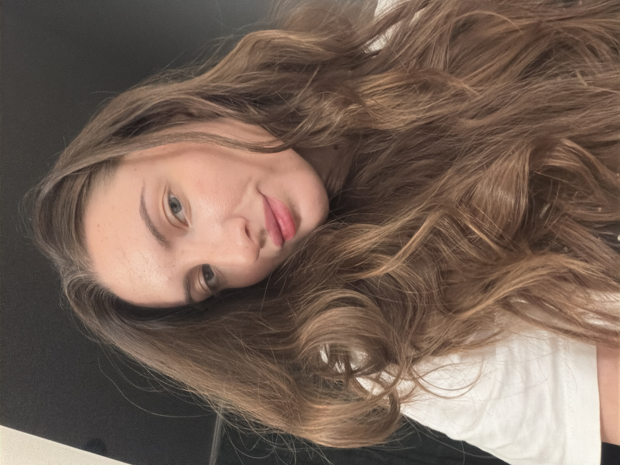 A close-up selfie of a woman with long, wavy brown hair is taken indoors. She is wearing a white t-shirt. The background is dark, with a lighter area on the left side of the frame. The image does not contain any information to suggest its location in Sharm El Sheikh, Egypt.