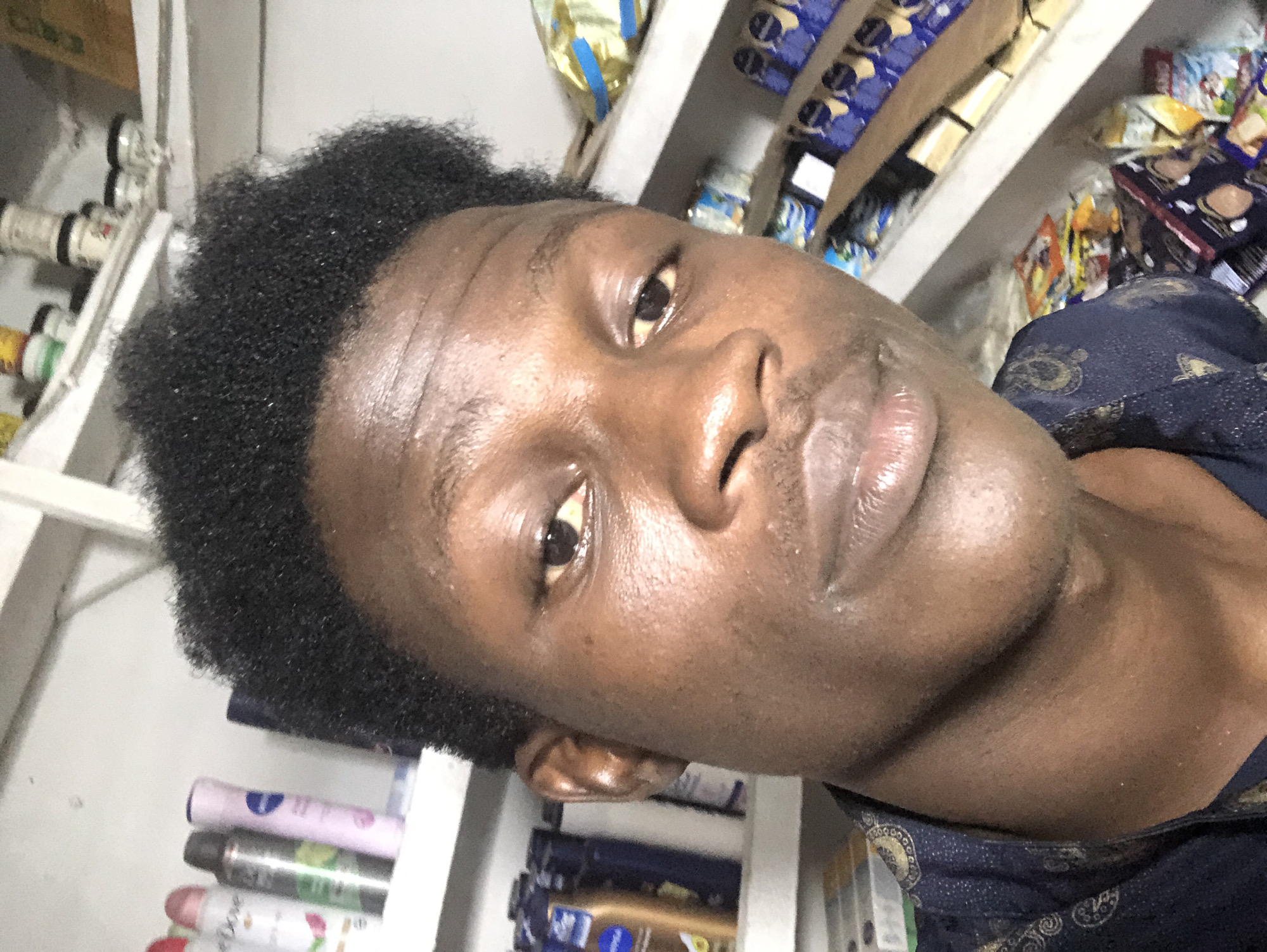 The image is a close-up selfie of a young Black man in a store. He is looking directly at the camera with a neutral expression. His hair is dark and textured, and he is wearing a dark blue patterned shirt. Behind him, shelves are stocked with various products, including personal care items and packaged goods. The lighting is bright, suggesting an indoor setting, likely a small shop or market. The environment appears to be organized with merchandise displayed on shelves. The man's skin tone is medium to dark brown, and he has a prominent jawline. The overall scene conveys a casual and everyday atmosphere within a retail space in Ado, Nigeria.