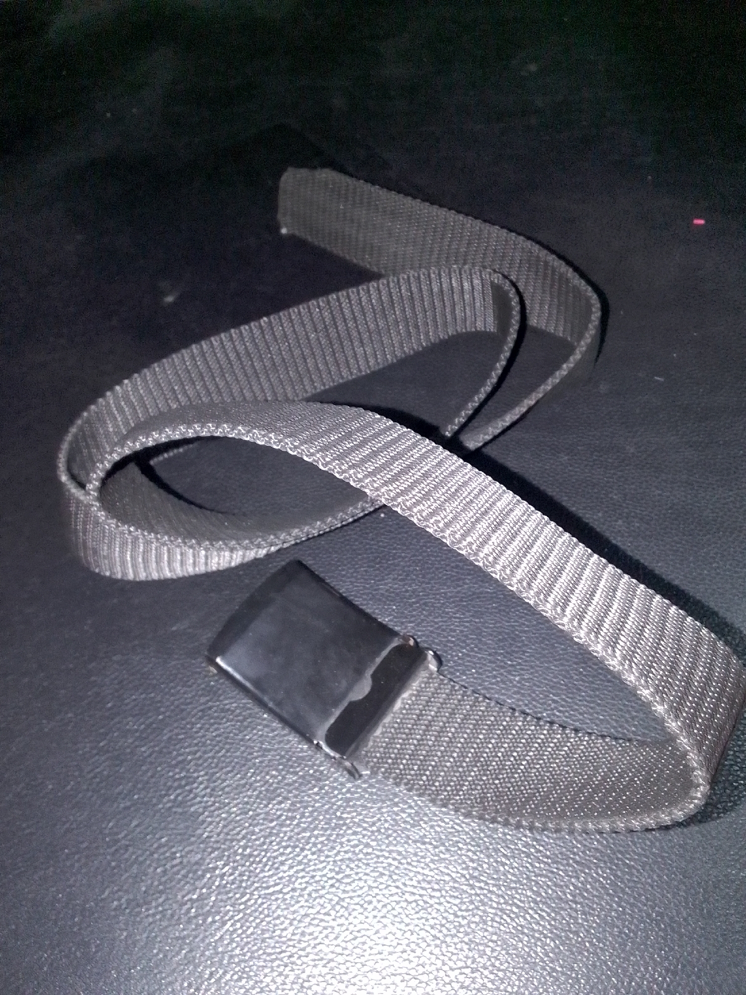 A coiled black fabric belt with a black plastic buckle lies on a dark, textured surface. The belt is positioned in a serpentine shape. The surface appears to be a dark material with a subtle grain, possibly leather or a similar synthetic. The lighting in the image is dim, creating shadows and highlighting the texture of the belt and the surface. The image does not provide any information about the location in Gunungmasigit, Indonesia.