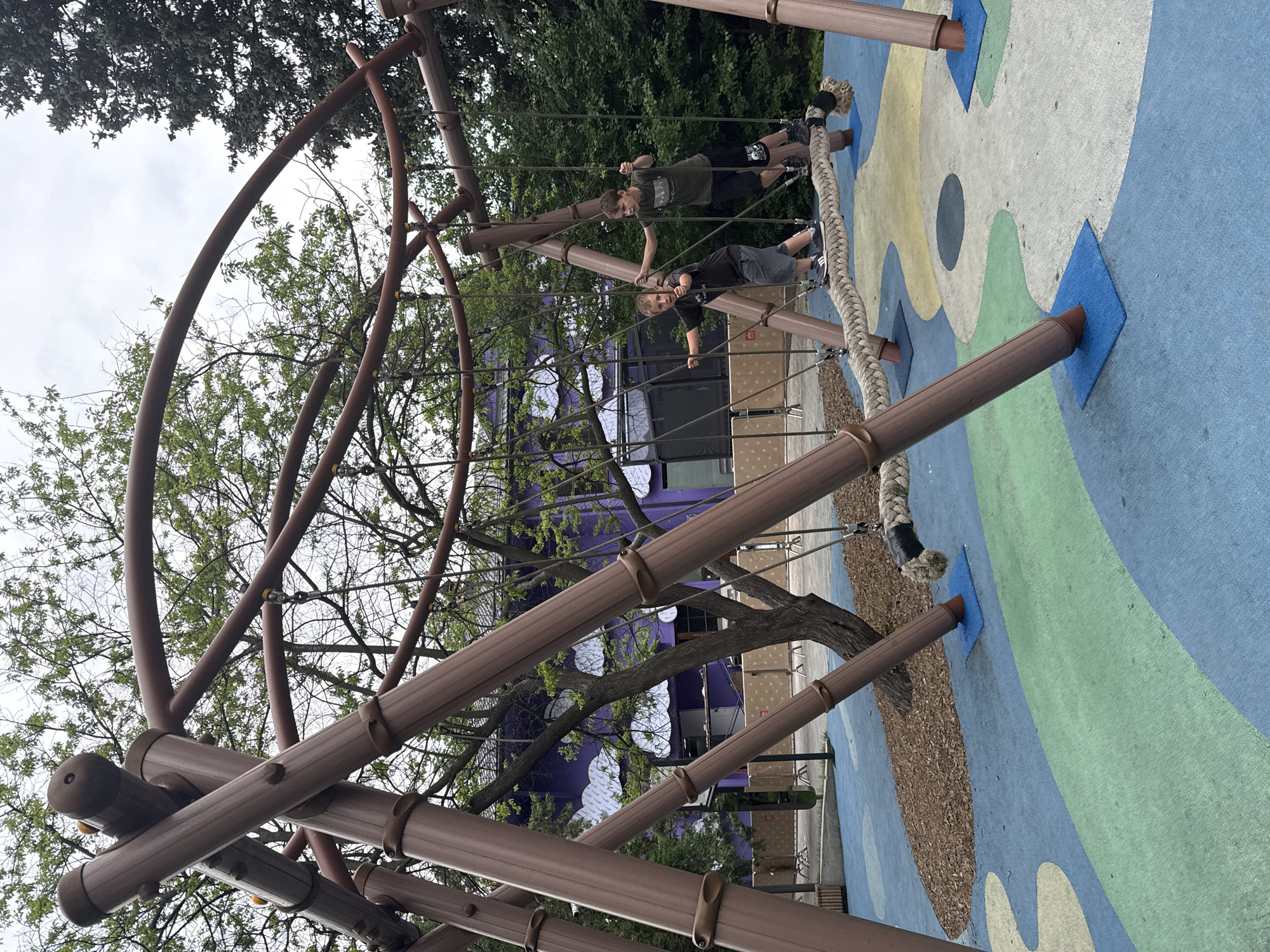Two young boys are playing on a rope swing apparatus in a playground. The structure is made of thick, brown, tubular supports and curved metal bars. The ground is covered in a colorful, soft-surfaced material with blue, green, and beige sections, and some areas of wood chips. In the background, there is a purple building with large windows and decorative white cloud-like shapes on its exterior. Trees with green foliage surround the playground. One boy, on the left, is wearing a dark t-shirt and grey shorts, and the other boy, on the right, is wearing a green t-shirt and black shorts. They are both holding onto the ropes of the swing. The sky is overcast. The location is not specified as Moscow, Russia.
