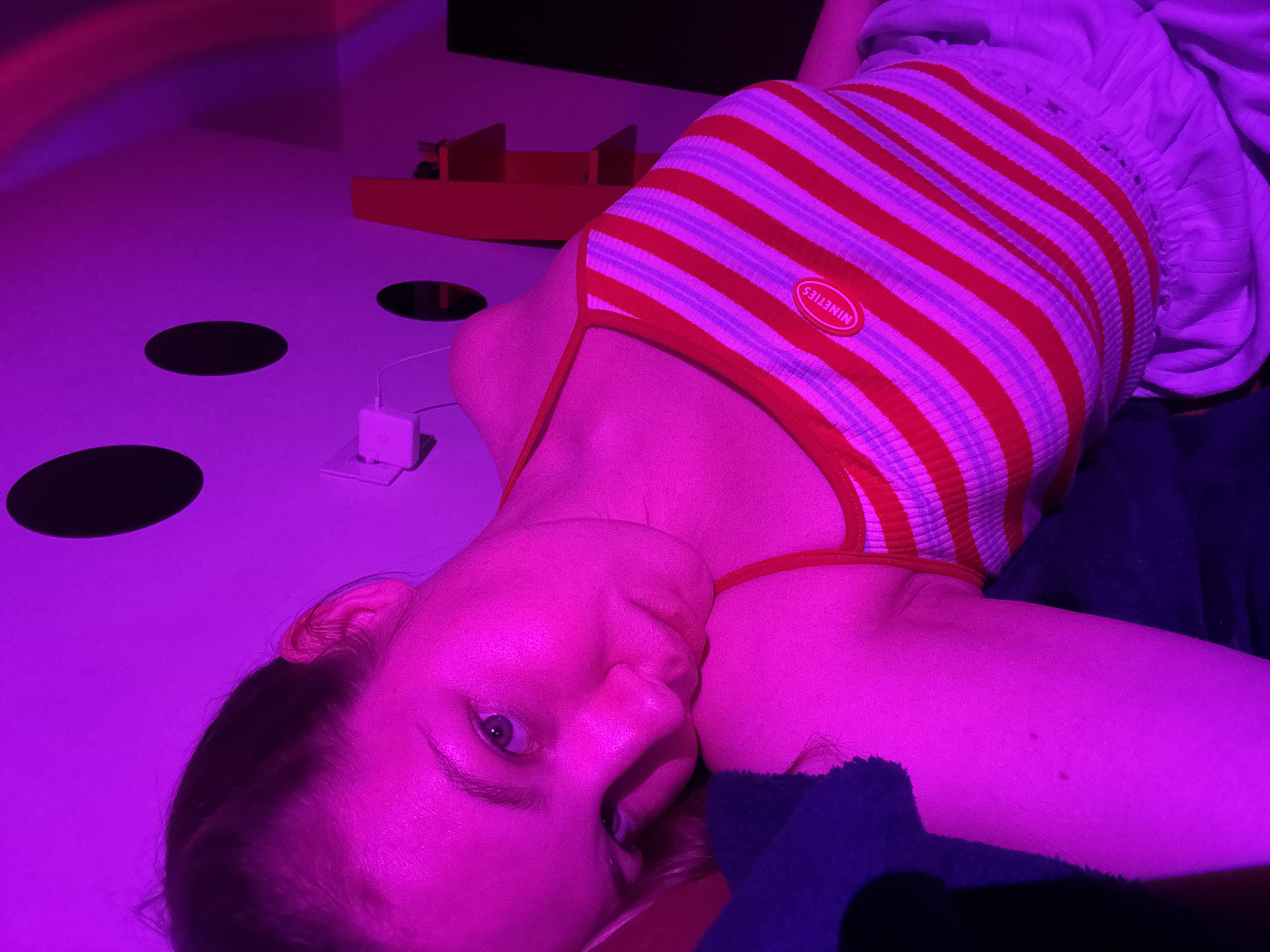The scene is an interior room within the Vozdovac Urban Municipality, Serbia, rendered under intense magenta-purple lighting. A young female subject is prominently featured, positioned in a reclining or resting posture, with her head leaning on her left arm/shoulder. She maintains direct eye contact with the viewer. Her dark hair is pulled back, and her complexion appears fair. She is dressed in a red and white horizontally striped, ribbed tank top with thin straps, featuring a red oval patch with the word "NINETIES" in white text on the upper left chest. Light-colored, loose-fitting shorts or pants are partially visible on her lower body. A dark blue or dark purple fabric is present beneath her left arm. The background consists of a light-colored wall adorned with several dark, circular decals. A white rectangular electronic device, possibly a Wi-Fi extender, is mounted on this wall with a connected cable. To the right of the subject, a reddish-orange shelving unit or similar furniture piece is visible. Faint bands of purple and red light, indicative of a projected light effect, are visible on the upper right wall/ceiling area.
