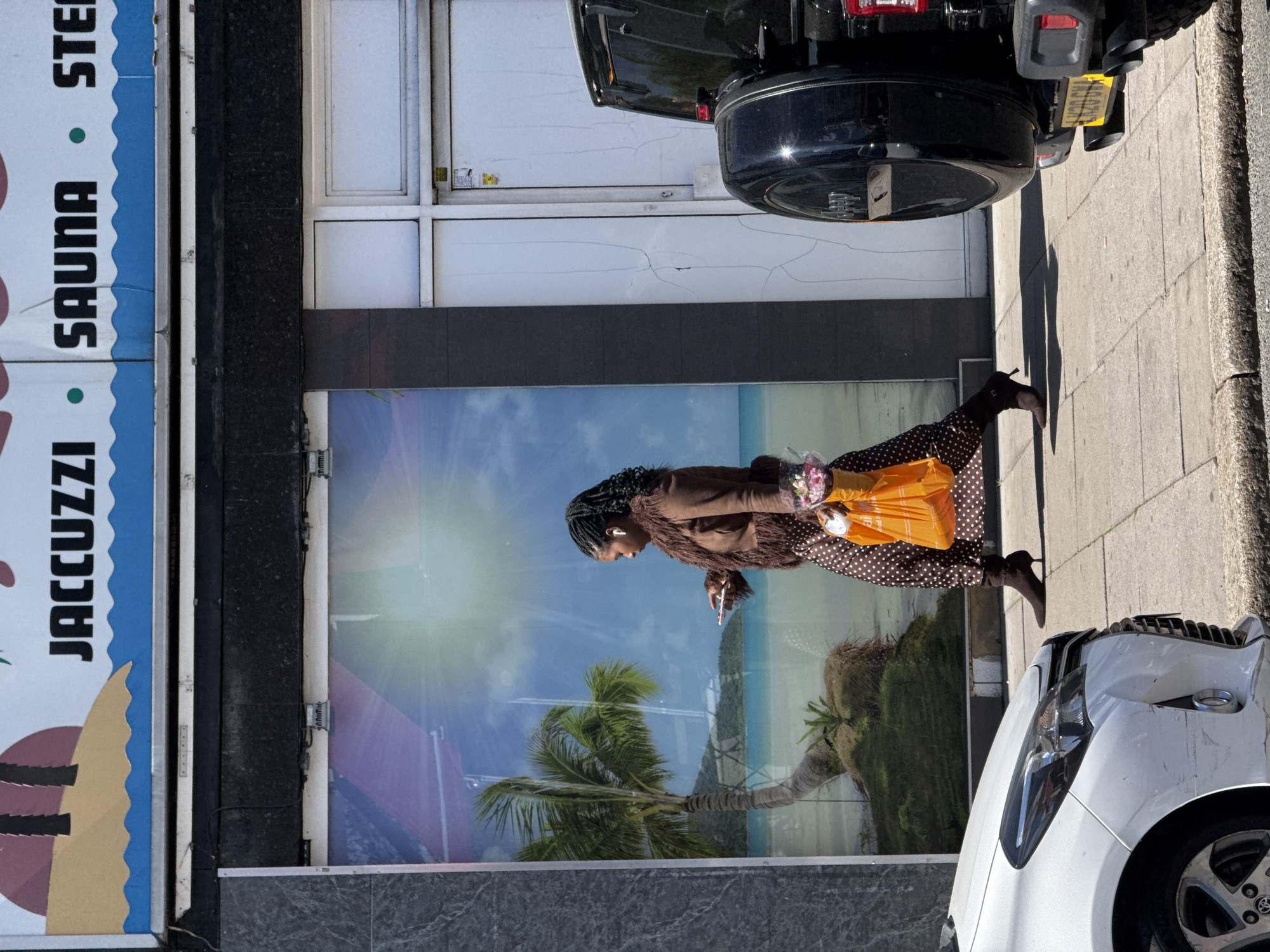 A woman is walking on a sidewalk in London, United Kingdom. She is carrying an orange shopping bag and a bouquet of flowers. She is wearing a brown jacket, a brown and white polka dot dress, and black high heels. The location appears to be a commercial district. To her left is a building featuring a poster for a spa, with the text "JACUZZI SAUNA STEAM". The windows on the building have an advertisement on them with a beach and palm tree on it. The woman appears to be looking at her phone or a similar electronic device. A white car is parked on the sidewalk near her. A black SUV is parked on the sidewalk behind her.