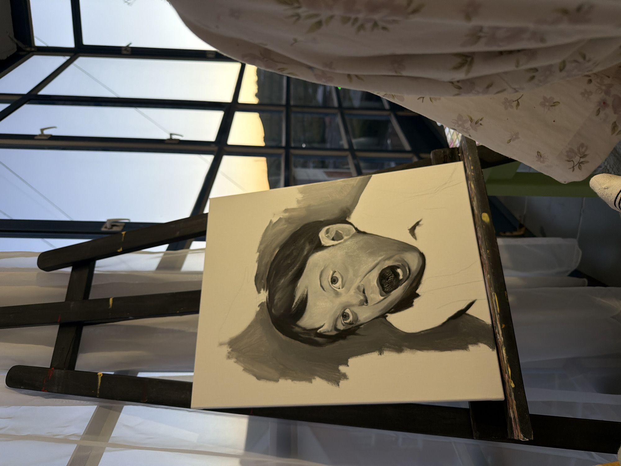 An easel holds a partially finished monochrome painting of a child's face with a wide-open mouth. The background of the painting is shaded dark gray. The easel is situated indoors, in front of a large window with a grid pattern. Through the window, a sunset or sunrise is visible over a cityscape with hills. A person, wearing a light-colored floral robe and white socks, stands to the right of the easel, partially obscuring the right side of the window. White curtains hang to the left of the easel. The scene appears to be a residential setting, possibly an apartment or house with a view.