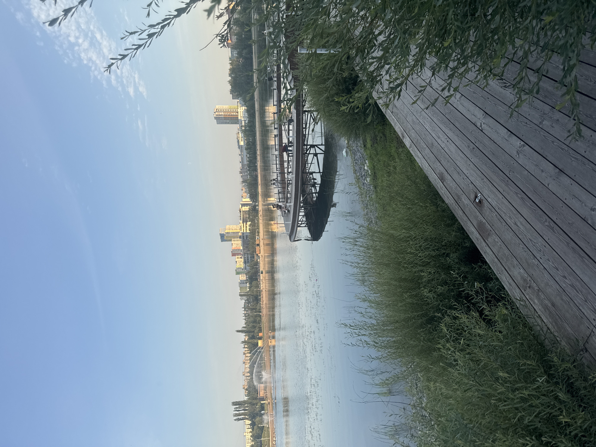The image depicts a waterfront scene in Ufa, Russia. In the foreground, a wooden boardwalk with a weathered texture extends diagonally from the bottom right corner. To the left of the boardwalk, lush green foliage borders the tranquil water. The water itself reflects the clear blue sky with scattered white clouds.
Across the water, a city skyline is visible, characterized by a mix of modern buildings and trees. A prominent feature in the middle ground is a circular wooden pier with benches, where several individuals are seated or standing. Further in the distance, a fountain sprays water into the air, adding a dynamic element to the landscape. The overall atmosphere is peaceful, suggesting an early morning or late afternoon setting.