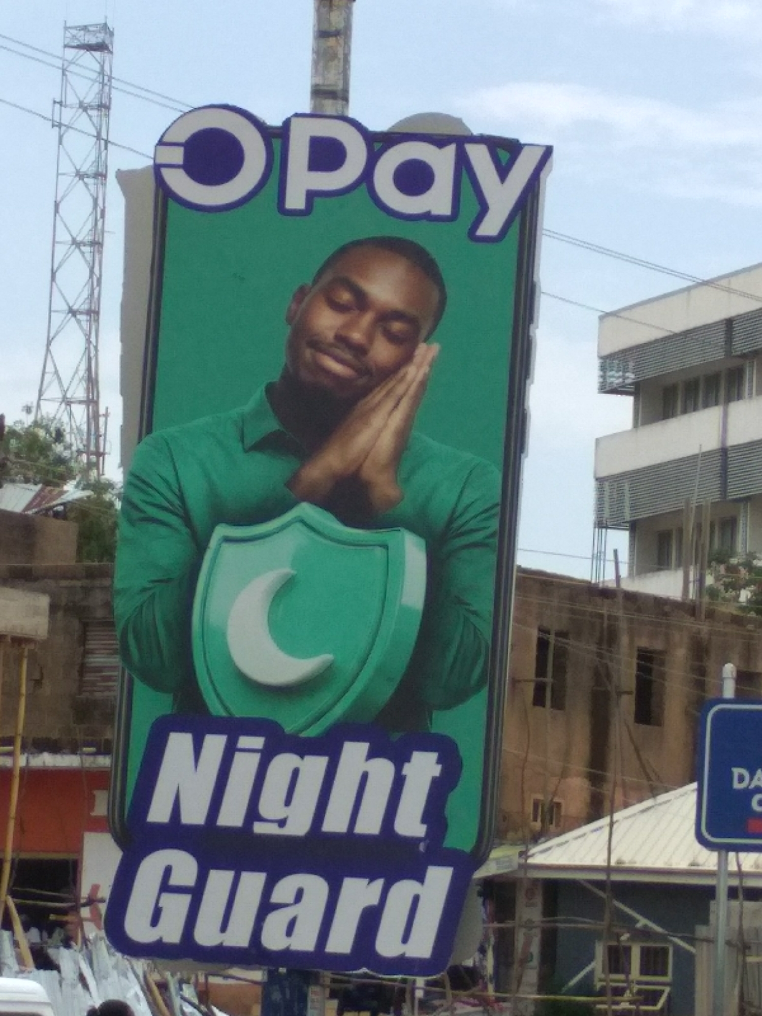 A tall, narrow advertising billboard for "OPay Night Guard" dominates the scene. The billboard features a man with dark skin wearing a green shirt, his hands clasped together against his face in a sleeping position. A green shield shape with a white crescent moon symbol is centered on his chest. The billboard is located outdoors in an urban environment, likely in Jos, Nigeria. Buildings are visible in the background, along with a communications tower on the left. A smaller blue and white street sign is partially visible on the lower right. The overall scene suggests a daytime shot in a bustling, developing urban area.