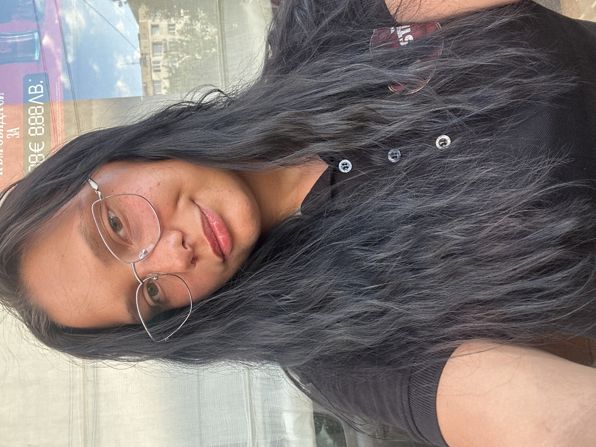 A medium shot depicts a woman with dark, wavy hair and wearing glasses. She is looking towards the camera, presenting a slight smile. She is wearing a black collared shirt with buttons. Behind her, visible through a window or glass surface, is a street scene. A sign with Cyrillic text and a Euro price is partially visible. The environment suggests an urban setting, possibly in Sofia, Bulgaria given the Cyrillic text. A building is visible in the background.