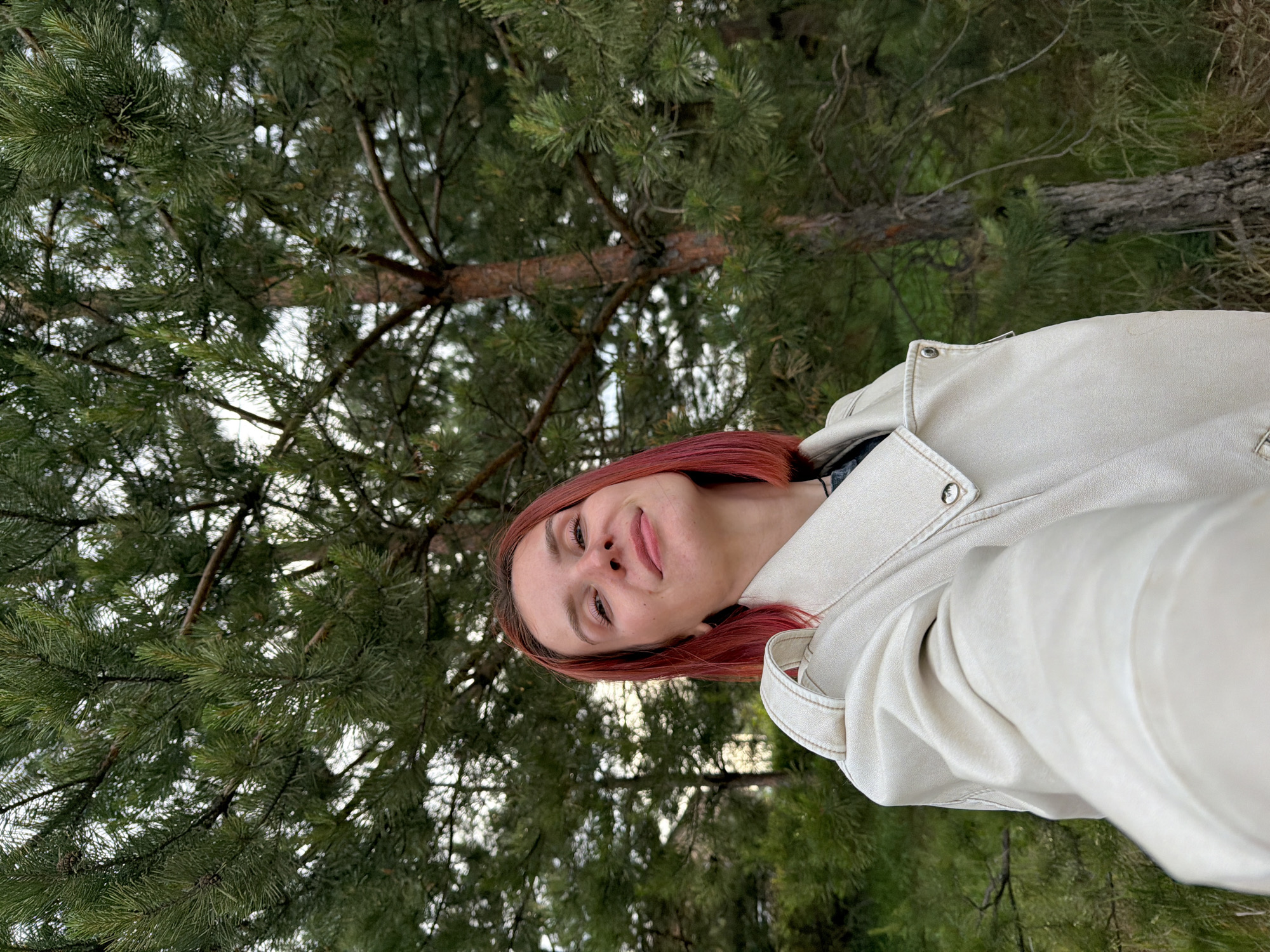 A woman with red hair wears a light-colored jacket and stands in front of a pine tree. The location is likely a park or forest in Chaikovsky, Russia.