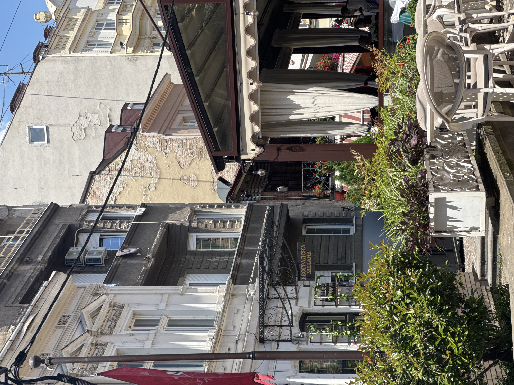 The image shows a street scene in Belgrade, Serbia, featuring a restaurant with outdoor seating. In the foreground, there are several white chairs and tables set up, some of which are covered by a glass-roofed awning. Lush green plants in decorative planters are positioned in front of the seating area.
Behind the restaurant, there are buildings of varying architectural styles and ages. The building on the left is a light-colored, ornate structure with large windows. Adjacent to it is a darker, more modern building with visible air conditioning units on a balcony. Further back, a building with peeling yellow plaster and exposed brickwork is visible, along with a lighter-colored building with a small square window and a decorative urn on its roof.
A red flag is partially visible on the left side of the image, attached to a lamppost. The signage above the entrance to one of the buildings reads "БЕОГРАДСКА МЕХАНА," which translates to "Belgrade Tavern." Several people are seated at tables inside the restaurant, enjoying the outdoor ambiance. The overall impression is a vibrant urban setting on a sunny day.