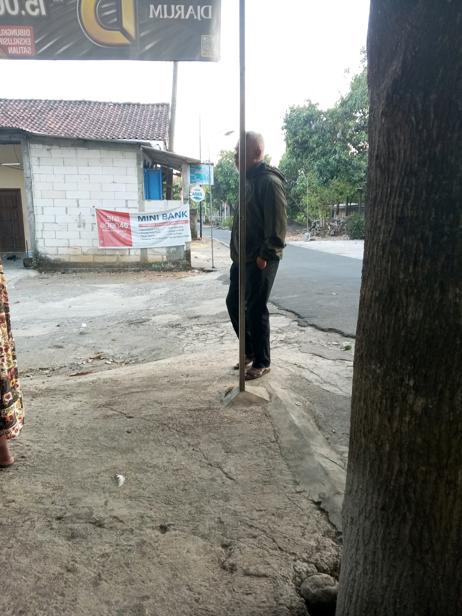 In Kedungringin, Indonesia, an outdoor scene shows a man standing next to a tree on a paved surface. The man, with short grey hair and wearing a dark green hooded jacket and black pants, stands with his hands in his pockets. He is facing to the right, with his back partially to the camera. In the background, a small white building with a red and white banner that reads "MINI BANK BNI agen46" is visible. The building has a tiled roof and a blue window. A road curves away into the distance, lined with trees and some debris. The ground is a mix of concrete and asphalt, with cracks and patches of dirt. A large tree trunk dominates the right side of the frame.