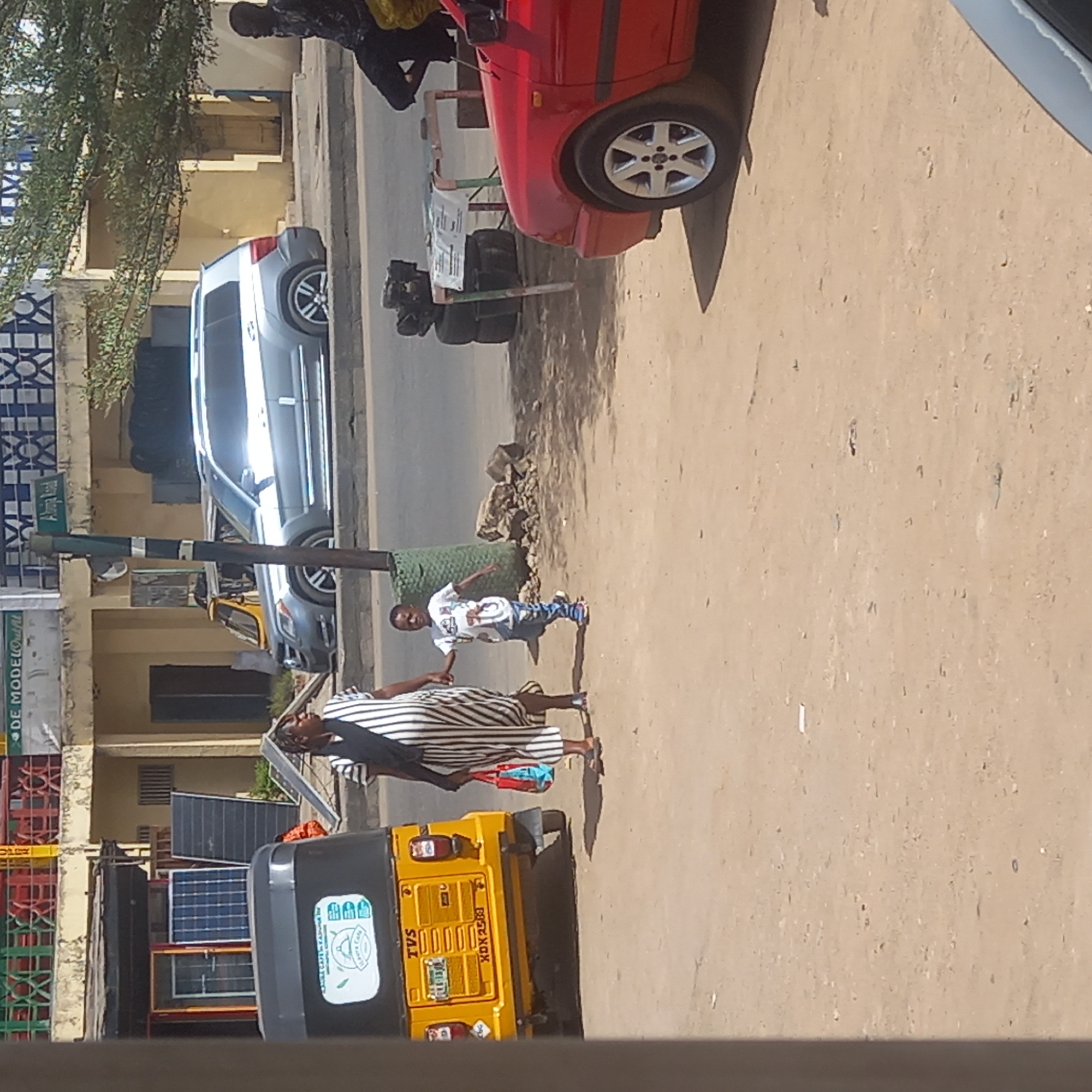 The image depicts a street scene in Sardauna, Nigeria. A yellow "keke" (tricycle taxi) sits on the right side of the frame. A woman wearing a black and white striped dress is walking with a young child near the keke. Further down the street, there are two parked cars (one silver and one red) on the left side. On the sidewalk, there's some construction material and debris near the cars. People are walking and standing near the parked cars. The environment appears to be a sunny day. Solar panels are visible beside the keke. A street sign is seen near the silver car.