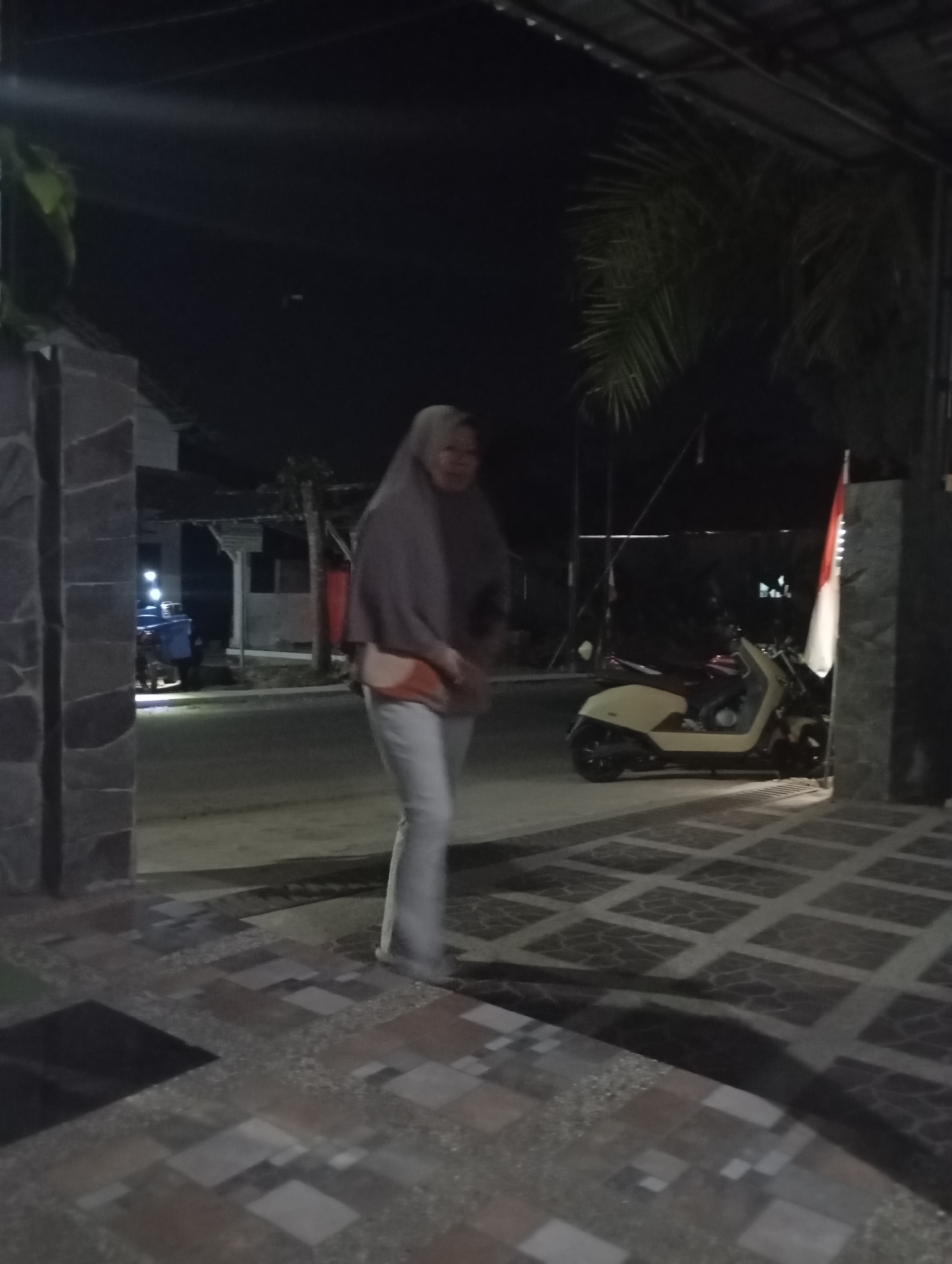 A woman in a light brown hijab and a light-colored outfit walks across a tiled area in front of a building at night. She is on the right side of the image, facing slightly away from the camera. Behind her, to her left, is a light-colored scooter. The scene appears to be outdoors, possibly a courtyard or walkway, with a patterned tiled floor and a paved road in the background. The building has a dark roof and some illumination coming from inside or from streetlights. A red and white flag is visible behind the scooter. The area appears to be a residential or commercial space in Banjarsari, Indonesia.