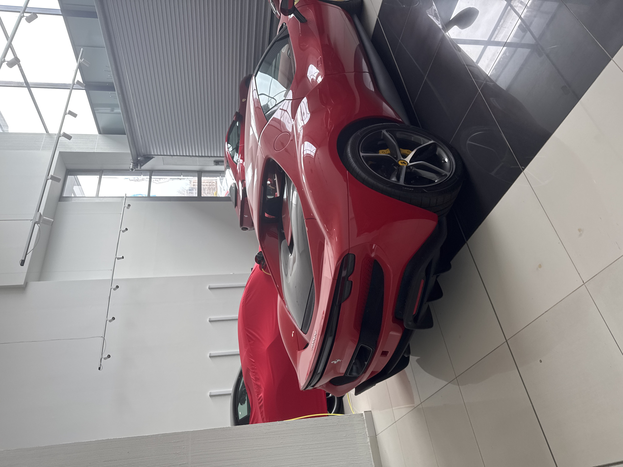 The image depicts the interior of a modern luxury automobile showroom or service center located in Sofia, Bulgaria. The central subject is a highly polished red Ferrari 296 GTB, positioned on the right side of the frame and viewed from its rear-three-quarter angle. Its black rear diffuser, dual central exhaust outlets, and five-spoke dark alloy wheels with yellow brake calipers are visible. To the left and slightly behind the 296 GTB, another red Ferrari coupe is partially visible, with its front angled away from the viewer. Further to the left, a third vehicle is completely obscured by a red car cover, with a yellow cable, possibly for charging, extending from its rear to the floor.
The flooring consists of large, glossy square tiles; white tiles cover the left side, transitioning to black tiles beneath the prominent Ferrari on the right. The walls are predominantly white and smooth, with subtle vertical structural or decorative elements present on the rear wall. Overhead, white track lighting fixtures with multiple spotlights illuminate the space. Large windows on the right side of the facility provide a view of an overcast exterior urban environment, revealing other buildings and faint greenery. A grey, horizontally slatted roll-up garage door is partially visible above and to the right of the cars. No individuals are present in the scene. The vehicles are stationary.