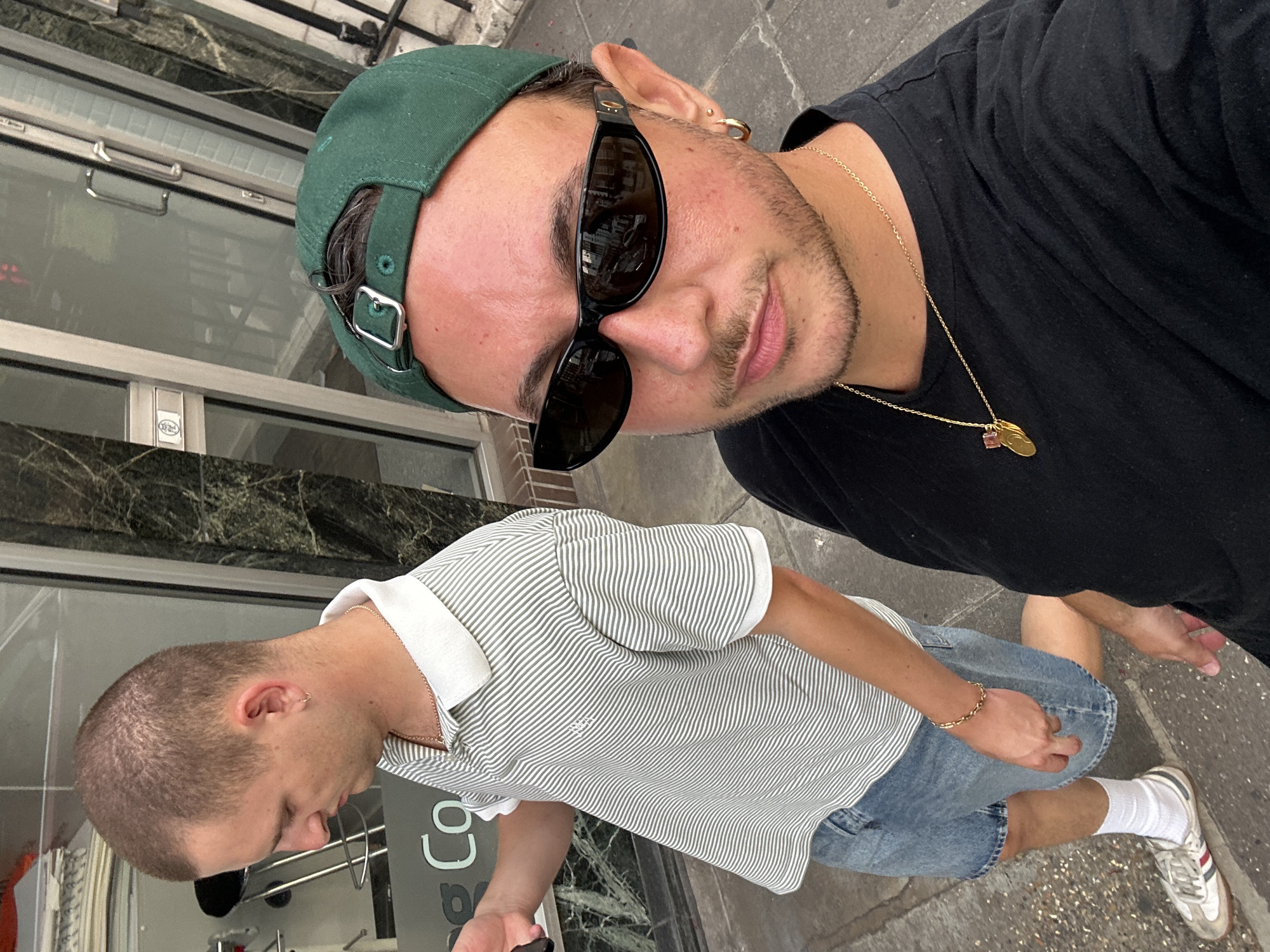 Two individuals are captured from the chest up in a street-level, outdoor photograph. The person in the foreground, facing the camera, is wearing a black t-shirt, a green baseball cap, and dark sunglasses. They have a gold chain with a pendant and a small hoop earring in their left ear. The person in the background, to the left, is seen from the side and is wearing a striped polo shirt, denim shorts, and white socks with athletic shoes. Both individuals appear to be walking. The background shows a building facade with large windows and a marble-patterned exterior. The scene is likely a street in London, United Kingdom, indicated by the architectural style and the general urban environment. The lighting suggests it is daytime.
