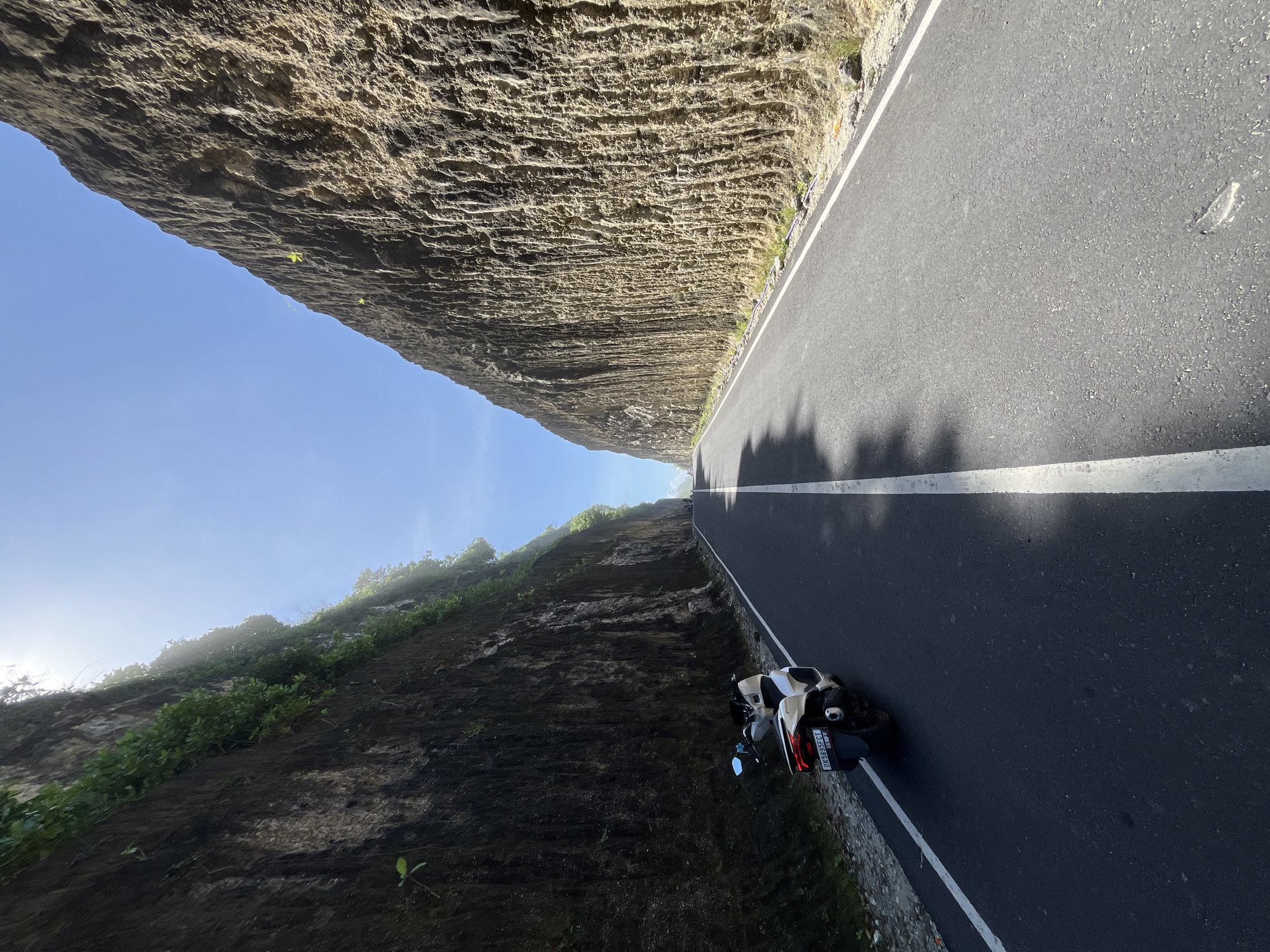 The image depicts a narrow asphalt road curving through a cliffside in Kuta Selatan, Indonesia. A white motorcycle is parked on the right edge of the road, facing away from the camera. The cliff on the left is tall and layered, with some vegetation at the base. The sky above is blue and slightly hazy. Shadows from foliage are cast on the road. The scene is brightly lit by sunlight.