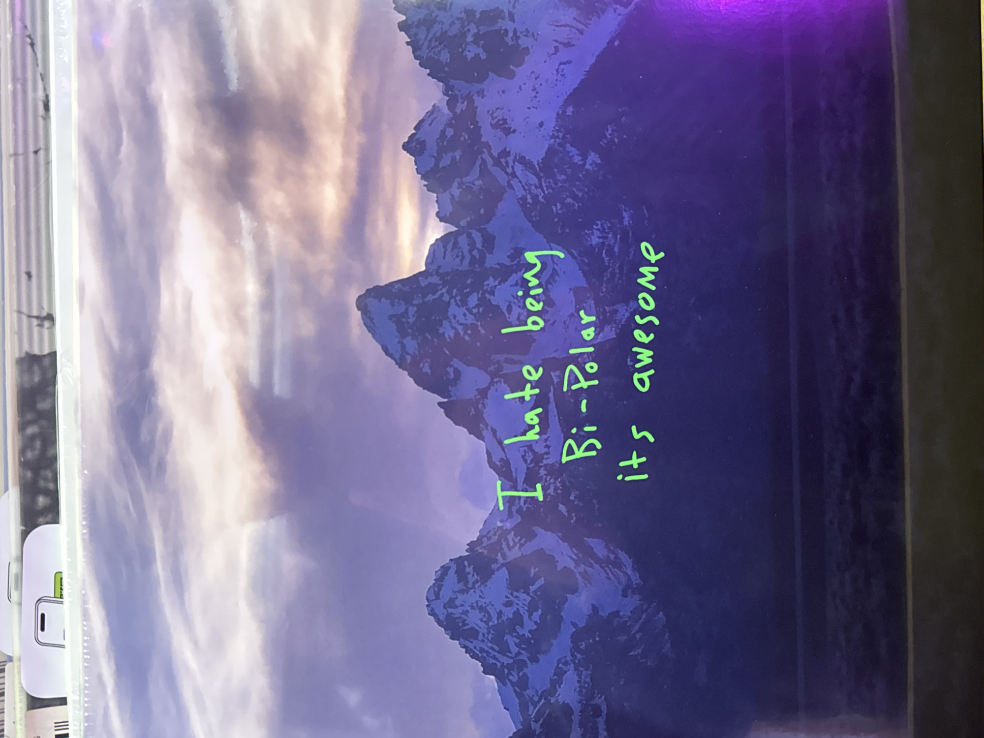 The image shows a vinyl record album cover. The cover features a landscape photograph of a mountain range with a cloudy sky. Superimposed on the mountain image in green marker is the text "I hate being Bi-Polar it's awesome". The album appears to be sealed in its original packaging. A barcode label is visible on the packaging in the lower left corner. The background is a dark surface, possibly a table or shelf. There is no indication of the location being Yerevan, Armenia.
