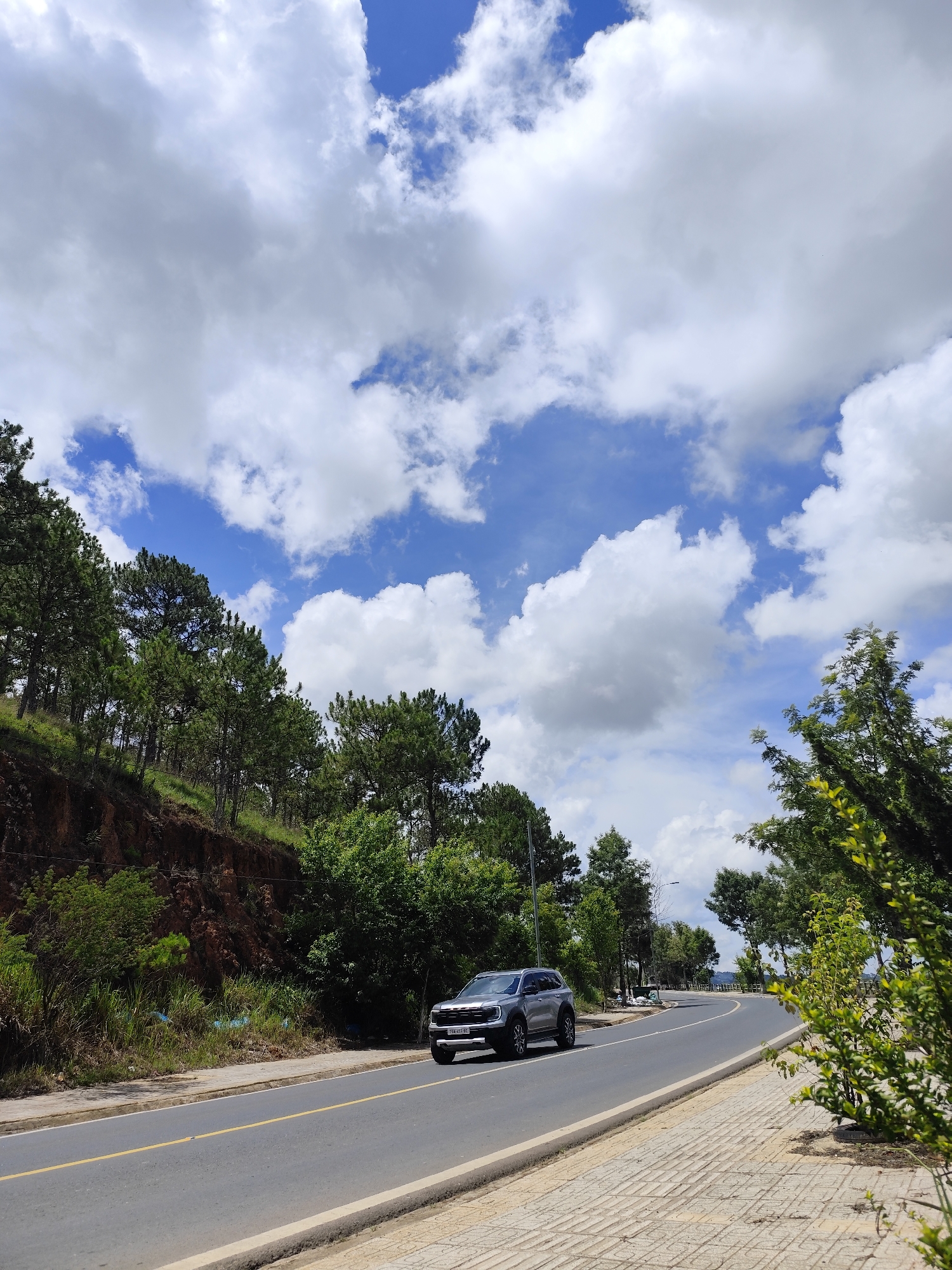 The image captures a sunny day in Lâm Viên - Đà Lạt, Vietnam. A grey SUV is traveling on a paved road. To the left, the road is bordered by a forested hillside with a mix of pine trees and other vegetation growing on a reddish-brown embankment. To the right, the road is lined with trees and a sidewalk paved with rectangular textured stones. The sky is partly cloudy with patches of blue sky visible between the white clouds.