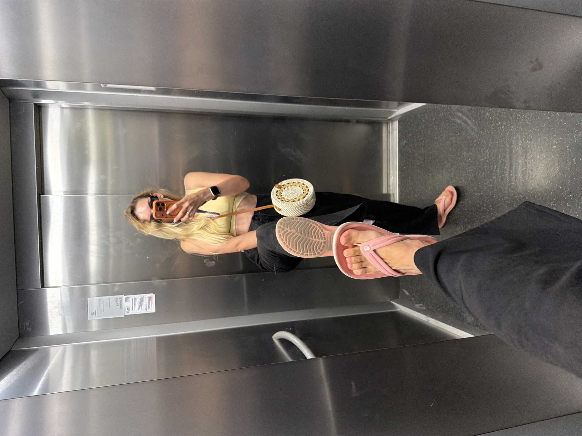 In Budva, Montenegro, a woman with blonde hair and sunglasses is taking a mirror selfie in an elevator. She is wearing a yellow crop top, black pants, and pink flip-flops. She is holding an orange-colored smartphone and has a round, woven straw bag. Another person's feet, wearing pink flip-flops and black pants, are in the foreground, suggesting the selfie is taken from their perspective. The elevator has stainless steel walls and a dark speckled floor.