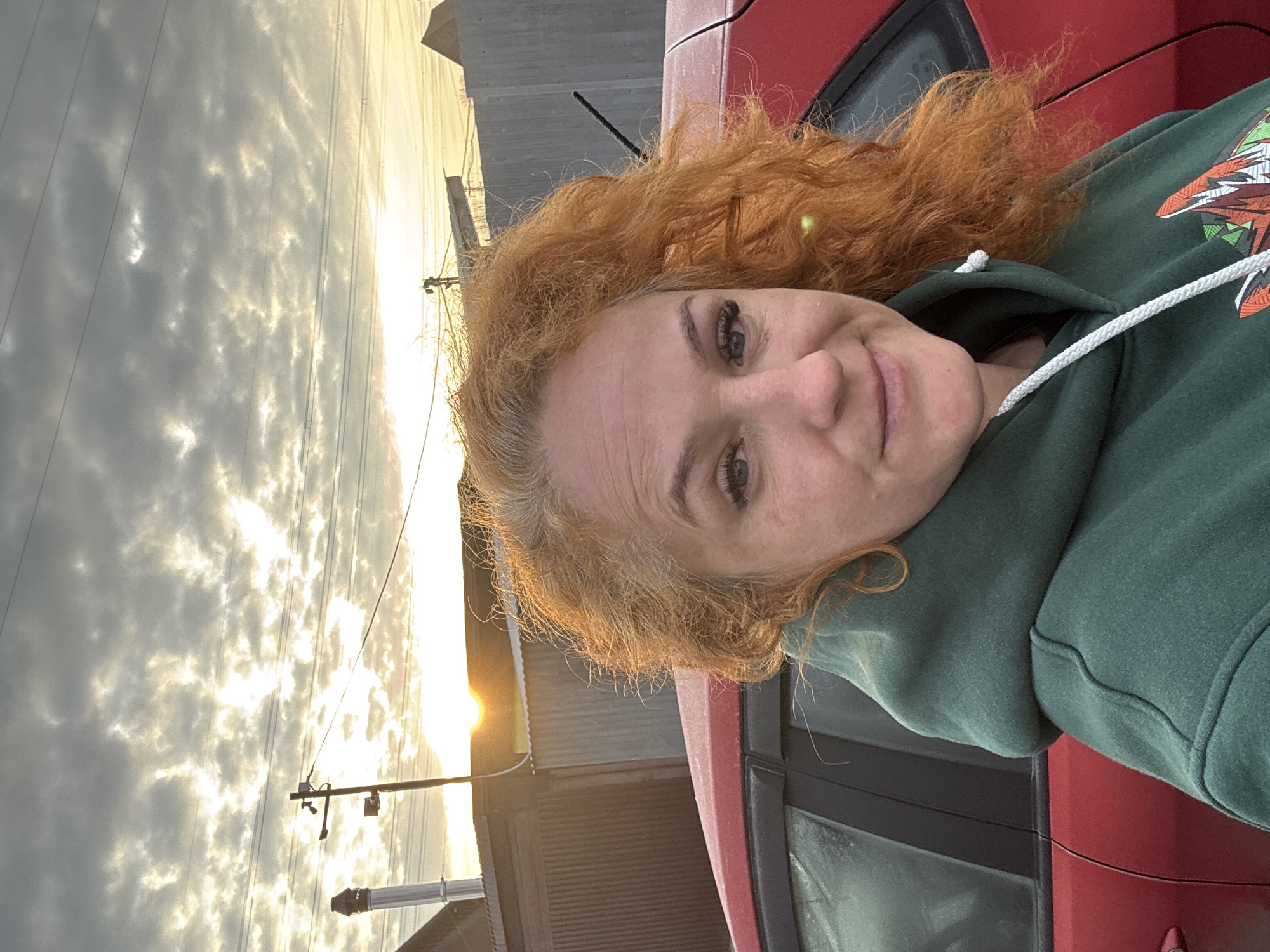 The image is a selfie of a woman with red hair, wearing a green hoodie. She is standing next to a red car. The background shows buildings and a sky with clouds and the sun setting. It could be in the city of Chaikovsky, Russia, based on the context. There is an electricity pole visible behind the building.