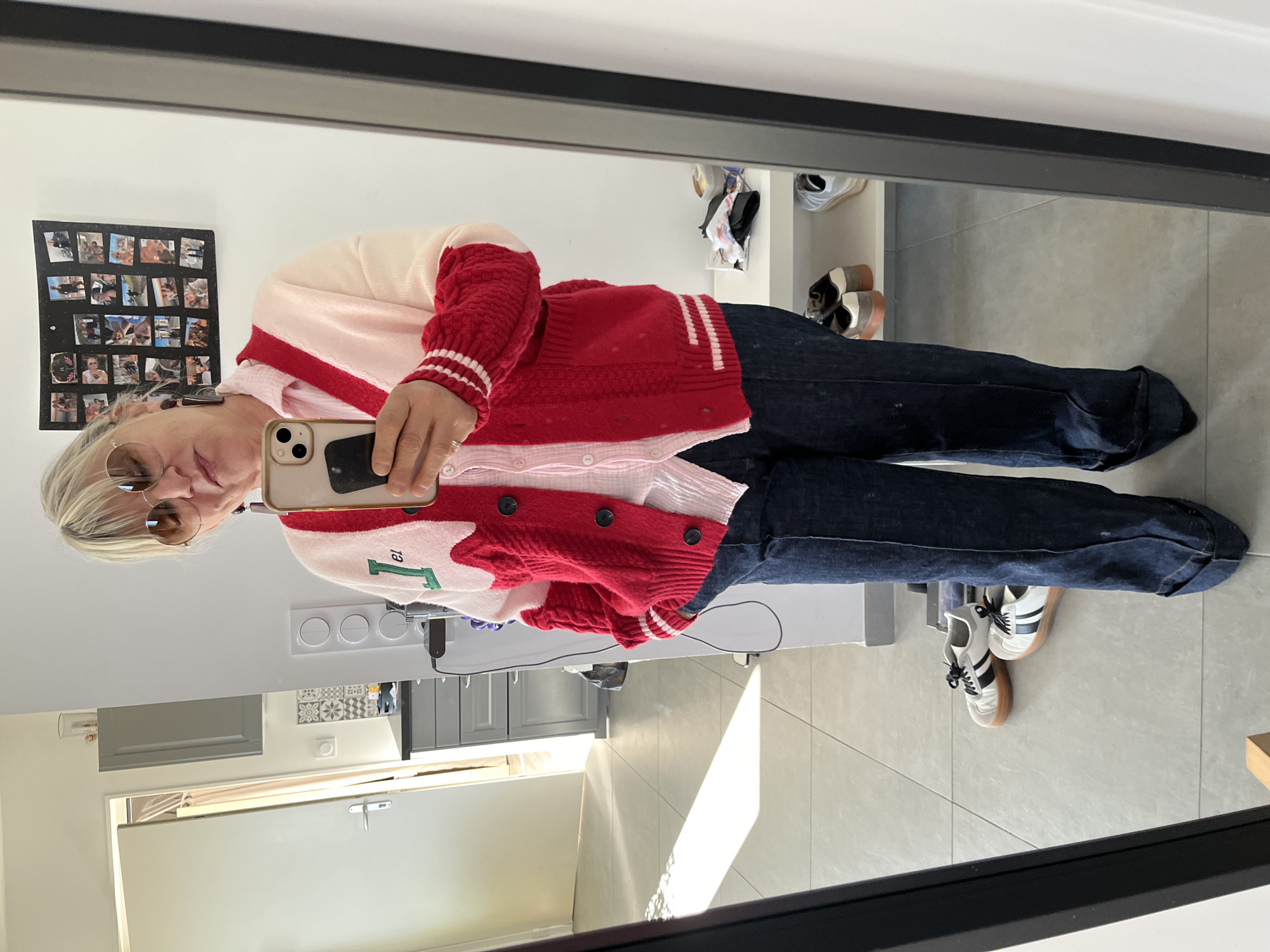 A woman is seen taking a selfie in a mirror indoors, in what appears to be a home. She is wearing sunglasses, a red and white cardigan, a pink shirt, and dark blue jeans. The location is Chambœuf, France. The background shows a glimpse of a hallway with a light-colored floor, gray kitchen cabinets, and a door. There are also shoes visible on a shelf and on the floor. A picture collage hangs on the wall behind the woman's head.