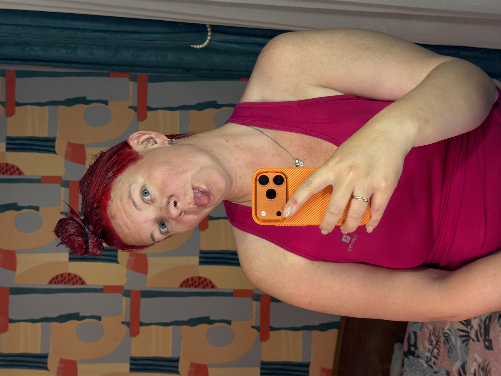 A woman with red hair, possibly in the process of being dyed given the residue on her scalp, is featured in a playful selfie. She is looking towards the camera with blue eyes, sticking her tongue out. Her hair is styled in a bun on the side of her head, and her left ear displays multiple piercings.
She is wearing a bright pink sleeveless top, with the inverted and mirrored text "SOMYOS" visible, likely a brand logo. A delicate silver necklace with a small charm hangs around her neck, and a silver-toned ring is on her right ring finger. She holds an orange smartphone with a textured case in her right hand, angled as if taking the photo.
The background is an indoor setting, featuring a patterned wall covering with abstract geometric shapes in muted tones of grey, orange, yellow, and dark blue. A dark teal curtain or fabric runs across the top of the frame. The scene is informal and lighthearted.
