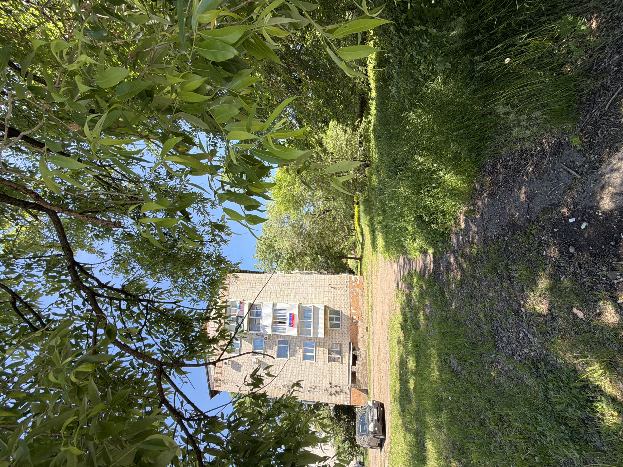 An outdoor, eye-level, full shot captures a multi-story apartment building with light-colored brick facade and a Russian flag hanging from a balcony. The building is situated in a grassy area with a dirt path leading towards it. A car is parked in front of the building. Trees with green foliage frame the scene, with some branches and leaves obscuring parts of the sky and building. The environment appears to be a residential area in Birobidzhan, Russia, with a clear blue sky overhead and dappled sunlight indicating a sunny day.