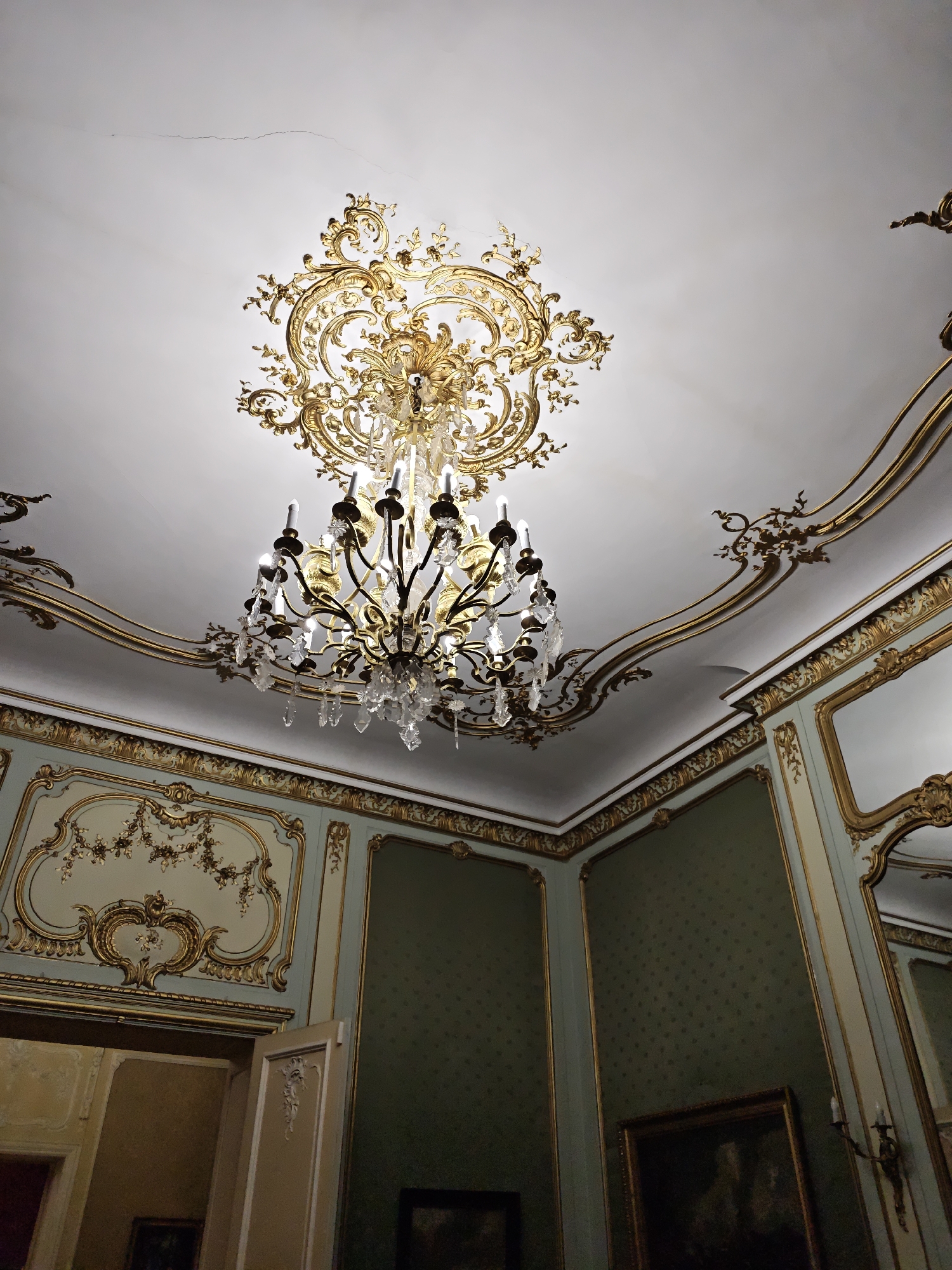 The image shows an ornate ceiling with a large, gilded chandelier and intricate gold stucco work. The walls are paneled with a light blue and gold decorative motif and green textured wallpaper. A doorway to the left reveals a dimly lit interior with paintings on the wall. The scene depicts a luxurious interior, likely a historical building. The chandelier is made of brass and crystal, with multiple arms holding faux candles. The ceiling has several cracks visible. The overall impression is one of opulent historical architecture. This image was taken in the Royal Hall of the Potocki Palace in Lviv, Ukraine.