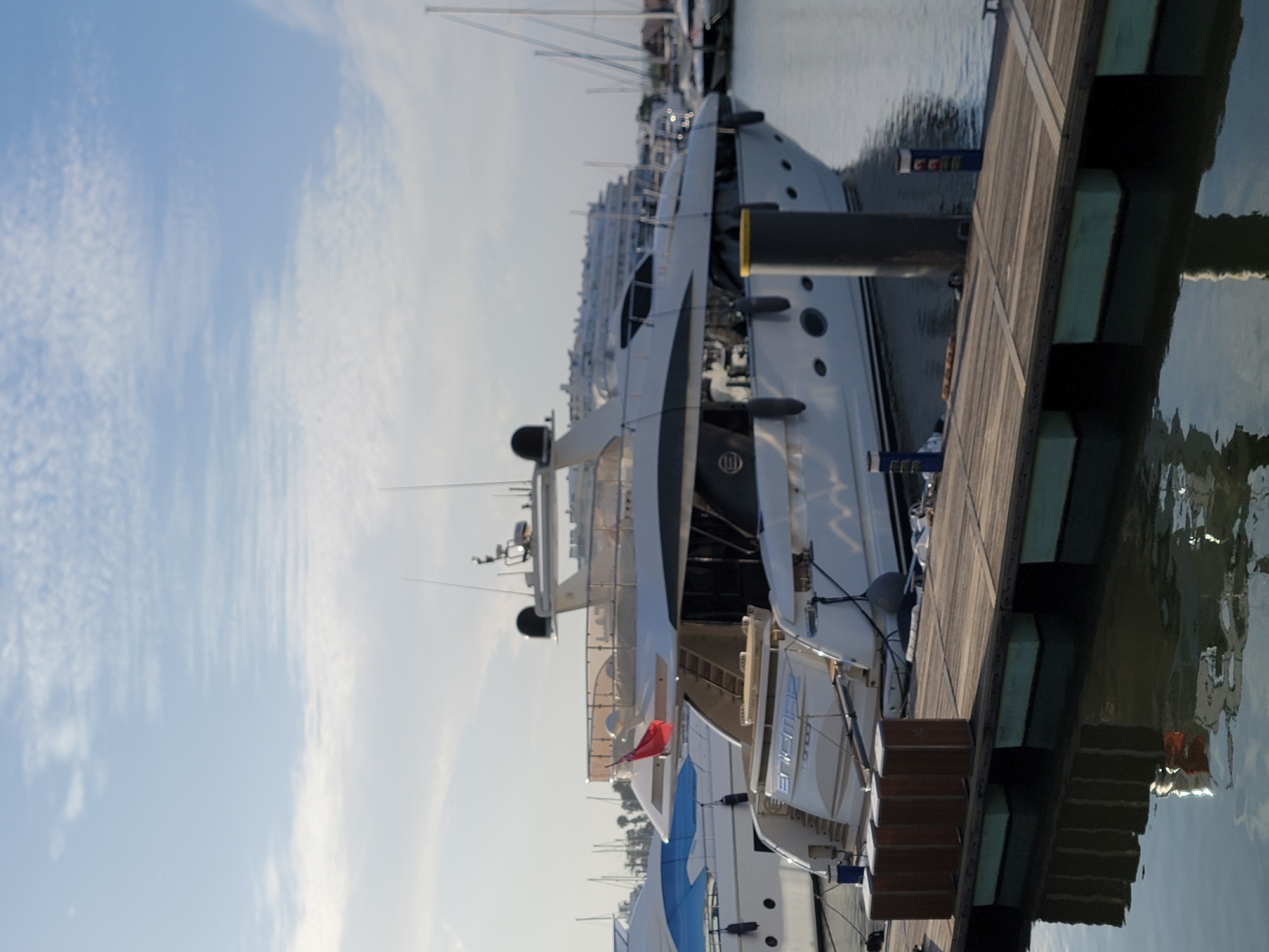 The image shows several large yachts docked in a marina. The scene is captured from a slightly elevated perspective, looking down at the yachts and the wooden boardwalk. The yachts are predominantly white with dark trim, and their sleek, modern designs are evident. One yacht, in the foreground, has a prominent nameplate that appears to read "ANASTASIA LONDON". A small red flag is visible on one of the yachts. The sky is a clear blue with scattered white clouds, suggesting fair weather. The sun is high in the sky, indicating it is likely daytime, possibly late morning or early afternoon. The water in the marina is calm, reflecting the sky and the yachts. The wooden boardwalk is constructed of weathered planks. In the background, other boats and masts are visible, along with what appears to be buildings or structures on the shore. The overall impression is one of a luxurious and tranquil marine environment. The location context provided is Quarteira, Portugal, which aligns with the presence of a marina and luxury yachts. There are no people visible in the image. The image has been rotated, and the subjects appear to be oriented vertically in the frame.
