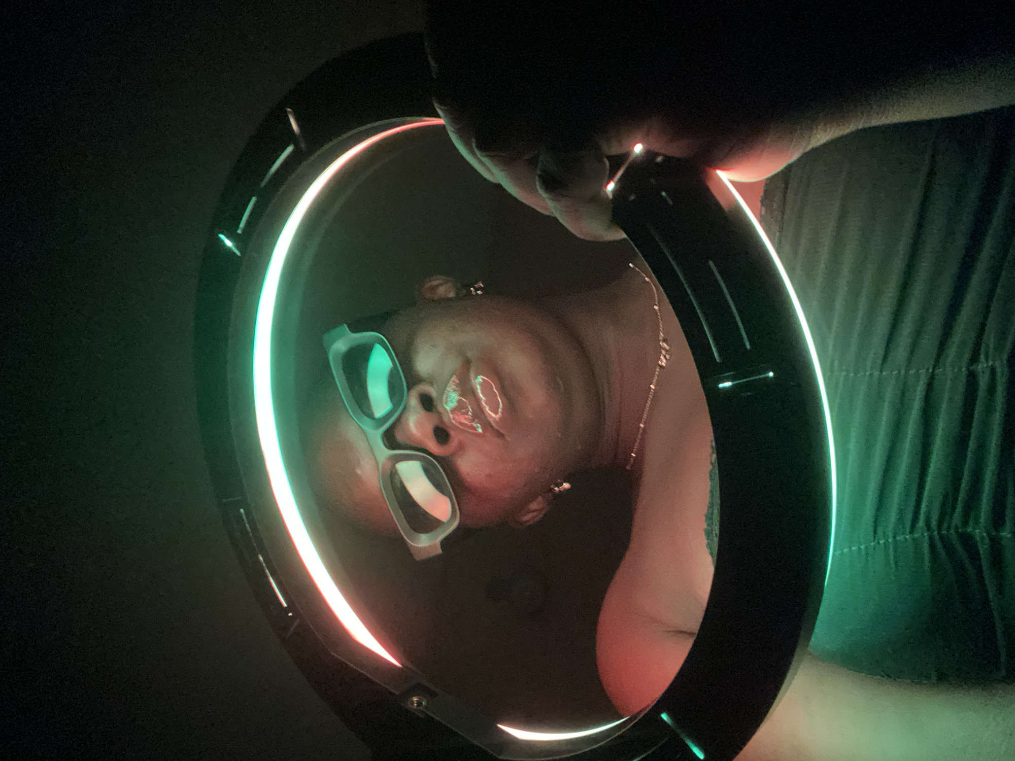 The image presents a stylized, close-up portrait of a person's face, seemingly reflected or positioned within a glowing, multi-colored ring light. The circular light emits vibrant hues of red, green, and white/cyan, providing the primary illumination for the scene. The person's face is inverted or significantly tilted, featuring prominent glossy lips and light-framed glasses. A delicate necklace with small charms is visible around the neck. A hand is seen grasping the top of the ring light, suggesting the device is being held up by the subject or another person. The background is largely dark, with hints of dark green, possibly draped fabric or clothing, visible on the right side. The overall ambiance suggests an indoor, low-light setting, likely at night, with the artificial light creating a dramatic and artistic effect. No visible text is present. The activity appears to be a self-portrait or a selfie captured using a ring light. The location context for this image is Maigizo, Nigeria.