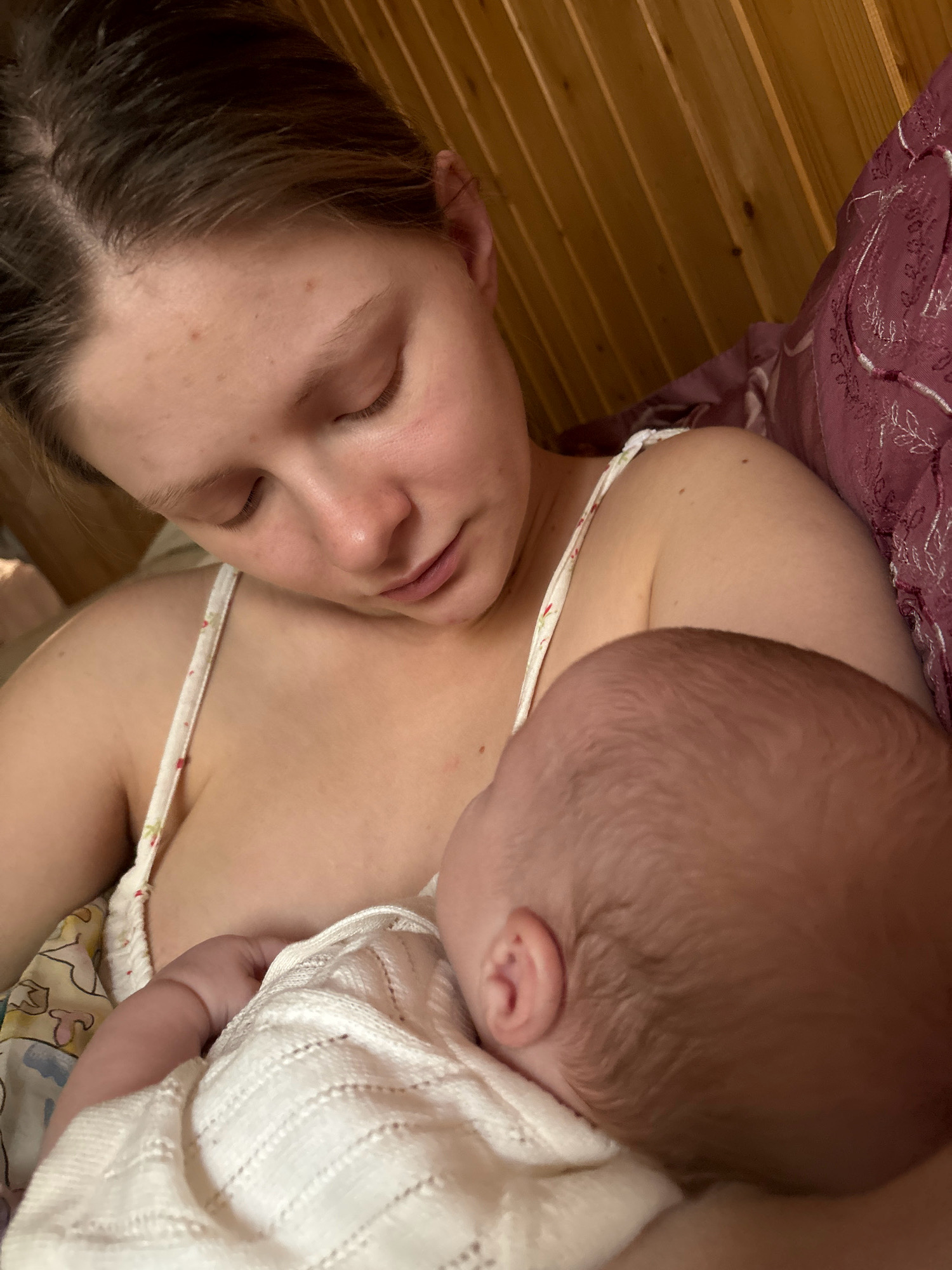 A young woman is lying down, holding a newborn baby to her chest. She is wearing a white tank top with a floral pattern. The baby is wrapped in a white knitted blanket and is being breastfed. The background consists of a wooden wall and a patterned fabric, possibly a quilt or bedspread. The scene appears to be indoors, in a bedroom. There is no visible indication of the location within the city of Взлет Снт, Russia.