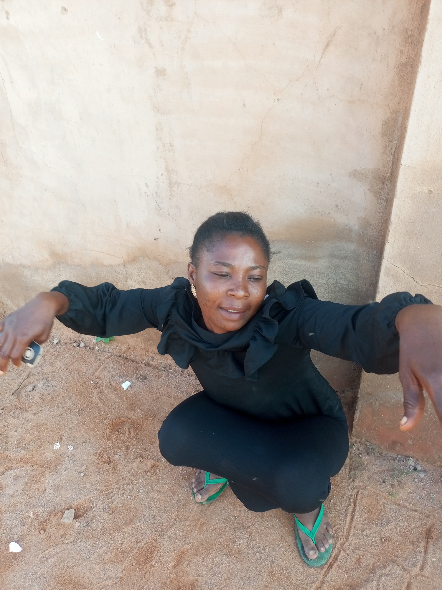 A woman is squatting on sandy ground in front of a light-colored, rough-textured wall. She is wearing a black dress with ruffled sleeves and black pants. On her feet, she has green flip-flops. She is holding a mobile phone in her left hand, and her right arm is extended towards the camera. Her eyes are closed, and she appears to be smiling. The environment suggests an outdoor location, possibly in a rural area or a less developed part of a city like Tempure, Nigeria, given the sandy terrain and simple wall construction.