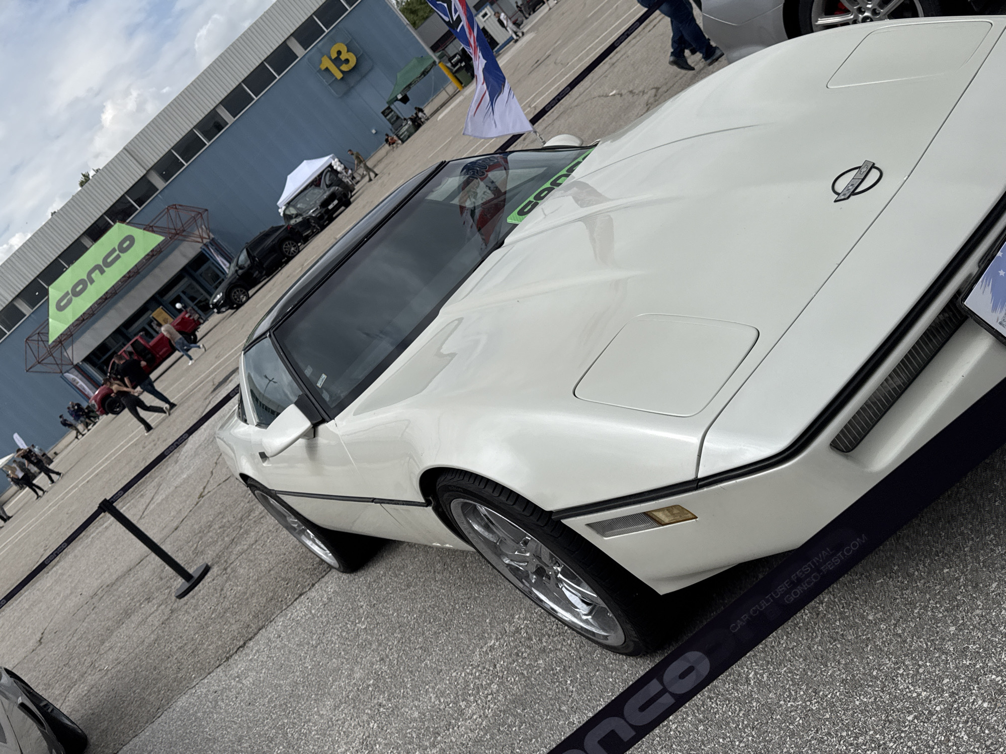 The image presents a white Chevrolet Corvette, likely from the C4 generation (1984-1996), parked at what appears to be an outdoor car event. The Corvette is the primary subject, positioned at an angle to the viewer, emphasizing its front and side profile. The car is clean and well-maintained, with chrome wheels and a black roof panel (T-top).
In the background, a building with the large number "13" is visible, along with a tent structure and other cars and people. A flag, possibly an Australian flag, is present near the Corvette. Black security stanchions surround the car. A banner with "CONCO CAR CULTURE FESTIVAL CONCO-FEST.COM" is situated near the car. The setting appears to be a paved outdoor lot, likely an exhibition ground or parking area.
While Plovdiv, Bulgaria, is referenced, there are no elements in the image itself to definitively confirm the location to be Plovdiv.