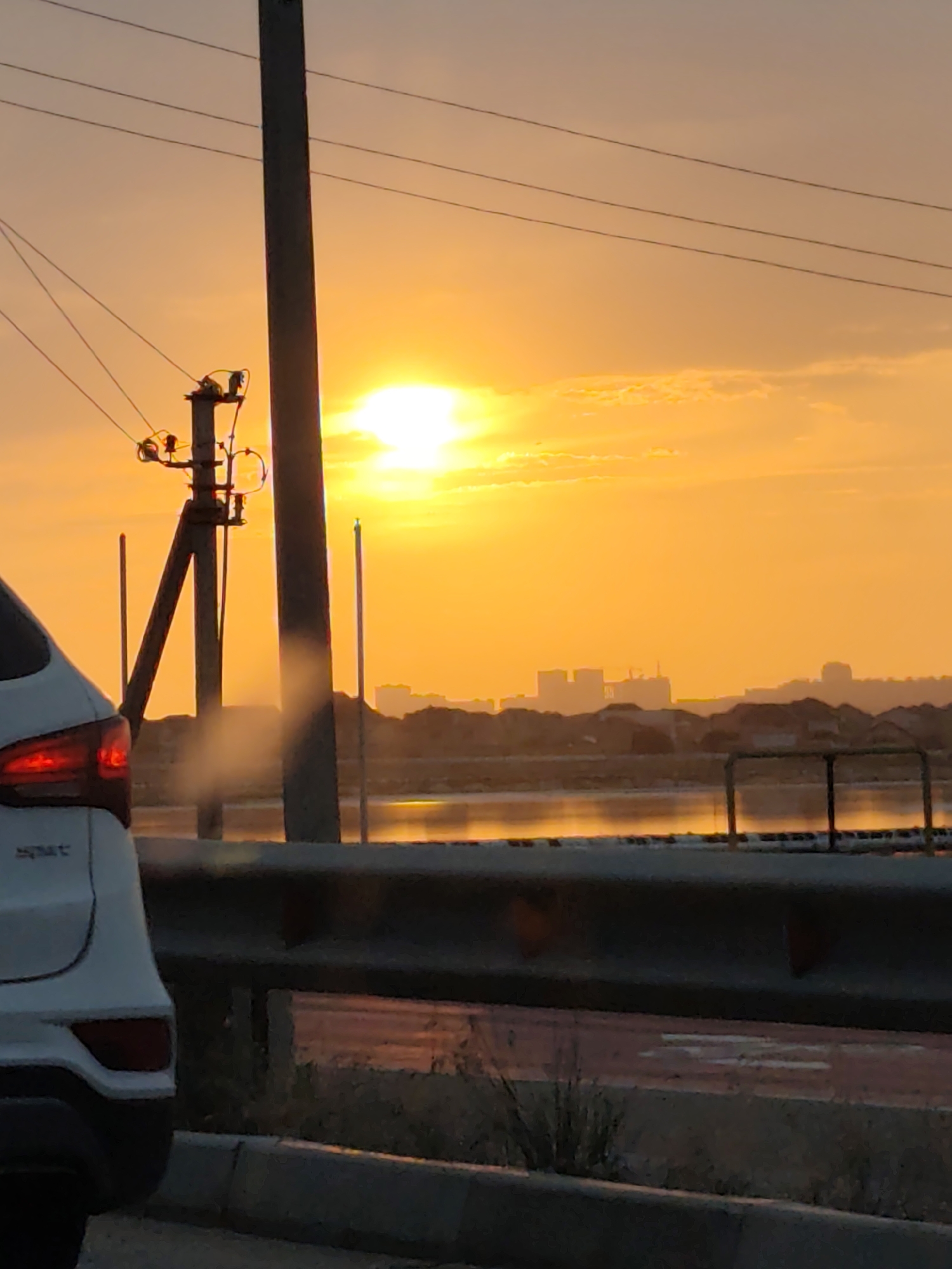 The image captures a vibrant sunset over Ömirzaq, Kazakhstan, with the sun a bright orange orb low in the sky, casting a warm glow across the scene. A white SUV is partially visible on the left, its taillight illuminated. Utility poles and power lines crisscross the sky, with one prominent pole in the foreground. In the distance, the silhouette of buildings and structures forms a hazy skyline. A body of water reflects the sunset's colors, and a barrier or fence is visible along its edge. The overall impression is a tranquil, expansive landscape bathed in the dramatic light of dusk.
