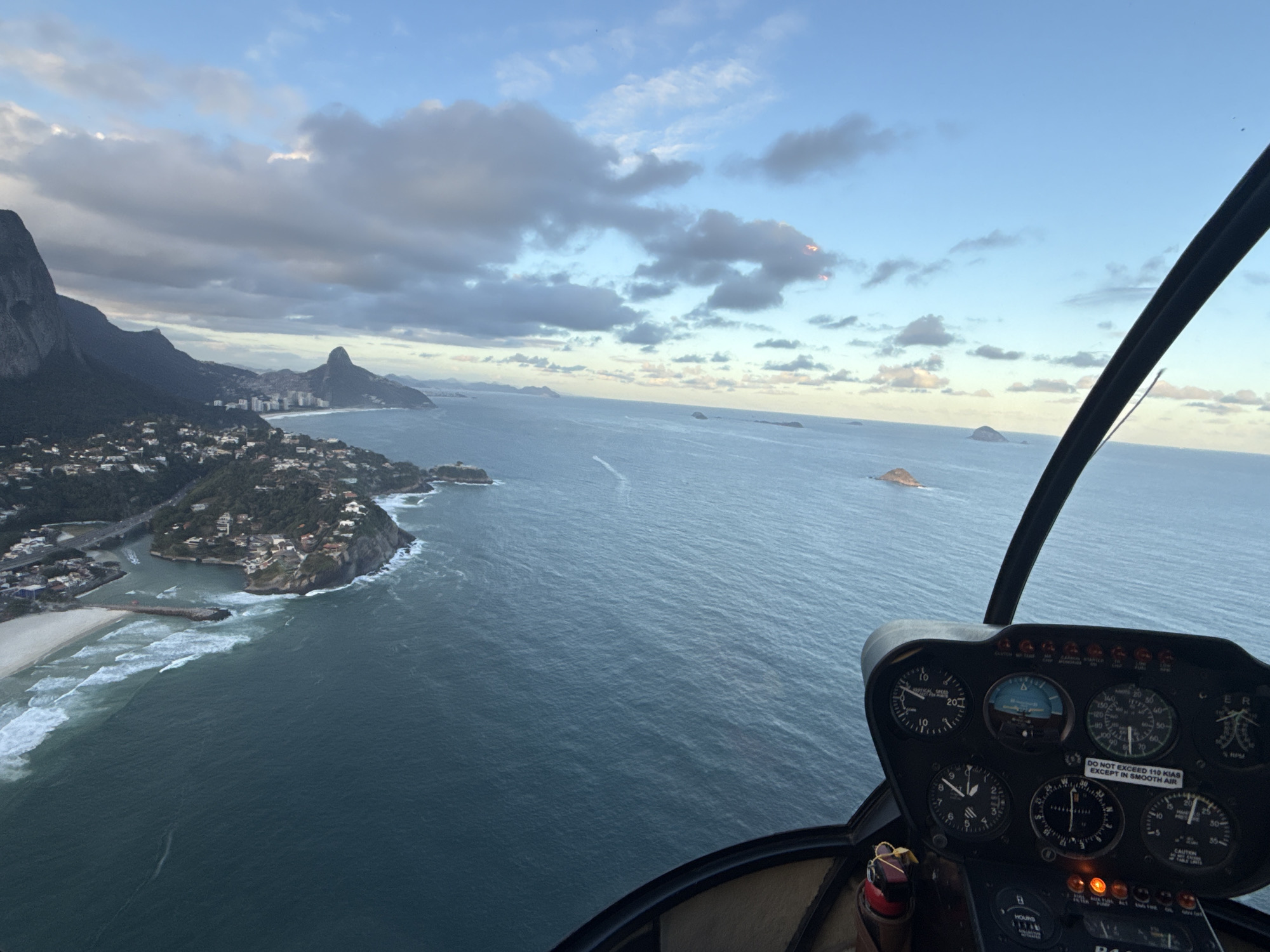 An aerial photograph, likely taken from a helicopter, captures a coastal urban landscape under a partly cloudy sky. In the immediate foreground, the helicopter's instrument panel is visible on the right, displaying various gauges, dials, and illuminated orange indicator lights. A warning label on the dashboard reads "DO NOT EXCEED 110 KIAS EXCEPT IN SMOOTH AIR." A black structural strut of the helicopter's frame extends diagonally along the right edge.
The primary scene depicts the coastline of Rio de Janeiro, Brazil, specifically looking east-southeast over São Conrado beach and the Joatinga peninsula. On the far left, the massive, rugged face of Pedra da Gávea dominates the landscape, covered in dark green vegetation. Below and to the right of Pedra da Gávea, a densely populated residential area with numerous houses and buildings climbs the hillsides and extends along the curving coastline.
Further along the coast, São Conrado beach is visible with white waves breaking on the sand. A river or canal (Canal da Joatinga) empties into the ocean, with a bridge crossing it and a prominent breakwater extending from the shore into the water. Beyond this, the Joatinga peninsula juts into the ocean, densely covered with houses and vegetation.
In the mid-ground, the distinctive conical peak of Morro Dois Irmãos rises prominently, with a more urbanized area, including taller buildings, nestled along the beachfront at its base, consistent with Leblon and Ipanema. The vast expanse of the Atlantic Ocean stretches towards the horizon, dotted with several small, rocky islands.
The sky is a mix of light blue and heavy, textured grey clouds, with subtle hints of warm light (possibly dawn or dusk) reflecting on the underside of some clouds. The overall lighting suggests either early morning or late afternoon. No people are discernible in the image.