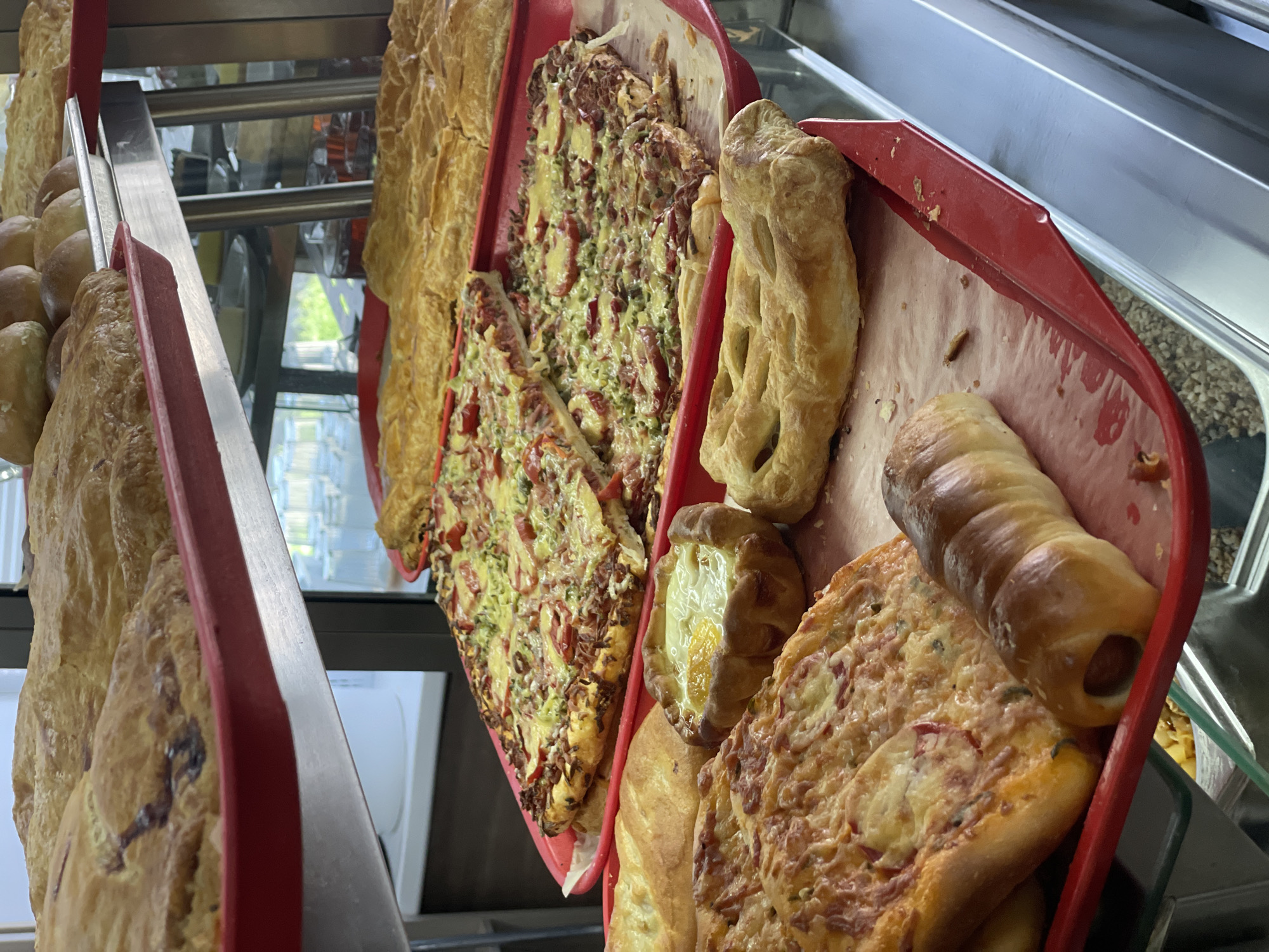 A close-up shot shows a display of baked goods, primarily in red trays, on shelves. In the top tray, large, golden-brown pastries with visible filling are arranged. Below this, a tray holds several square-shaped pizzas topped with melted cheese, vegetables, and meat. To the right and slightly lower, another tray contains large, flat, golden pastries. In the foreground, a red tray displays a variety of items: on the left, a square slice of pizza with cheese and toppings; in the center, a small pastry with a bright orange egg yolk visible within a baked crust; and on the right, a long, browned pastry roll, possibly containing a sausage. The background shows glimpses of more baked goods on higher shelves and a reflective surface, suggesting a food service environment like a bakery or cafeteria. The lighting is even, highlighting the textures and colors of the food.