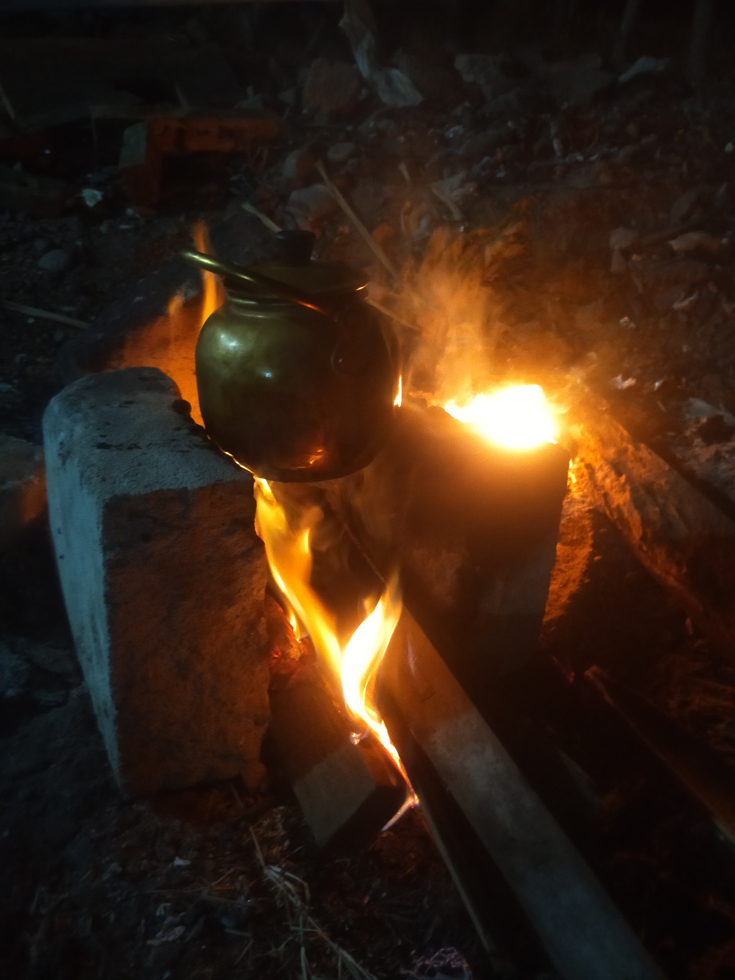In Pagerbarang, Indonesia, a brass kettle is being heated over an open fire in a dimly lit outdoor setting. The fire is supported by what appear to be two concrete blocks and some wooden logs. Flames are visible, and steam or smoke is rising from the kettle. The surrounding environment is dark and appears to be rough terrain with debris. The scene suggests a rustic or improvised cooking setup.