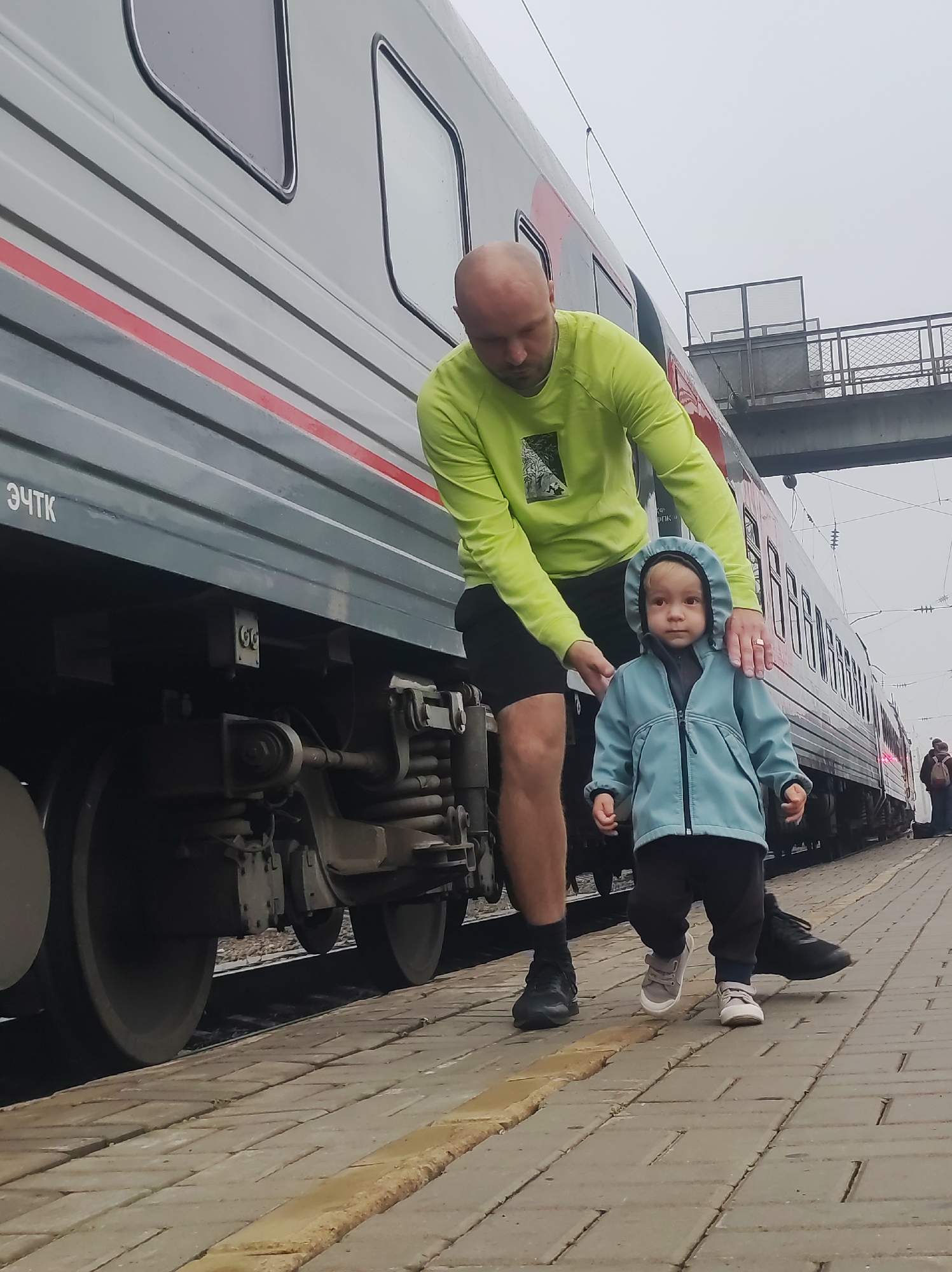 The image captures a father and his young child on a train platform in Kirov, Russia. The father, a bald man wearing a lime green long-sleeved shirt and black shorts, is leaning down to assist his toddler. The child, dressed in a light blue hooded jacket and dark pants, is walking forward, looking towards the camera. A train with gray carriages and a red stripe runs along the platform. The platform itself is paved with light-colored rectangular bricks. In the background, a pedestrian overpass is visible, along with other individuals and luggage, suggesting a busy transit area. The sky is overcast, indicating a cloudy day.