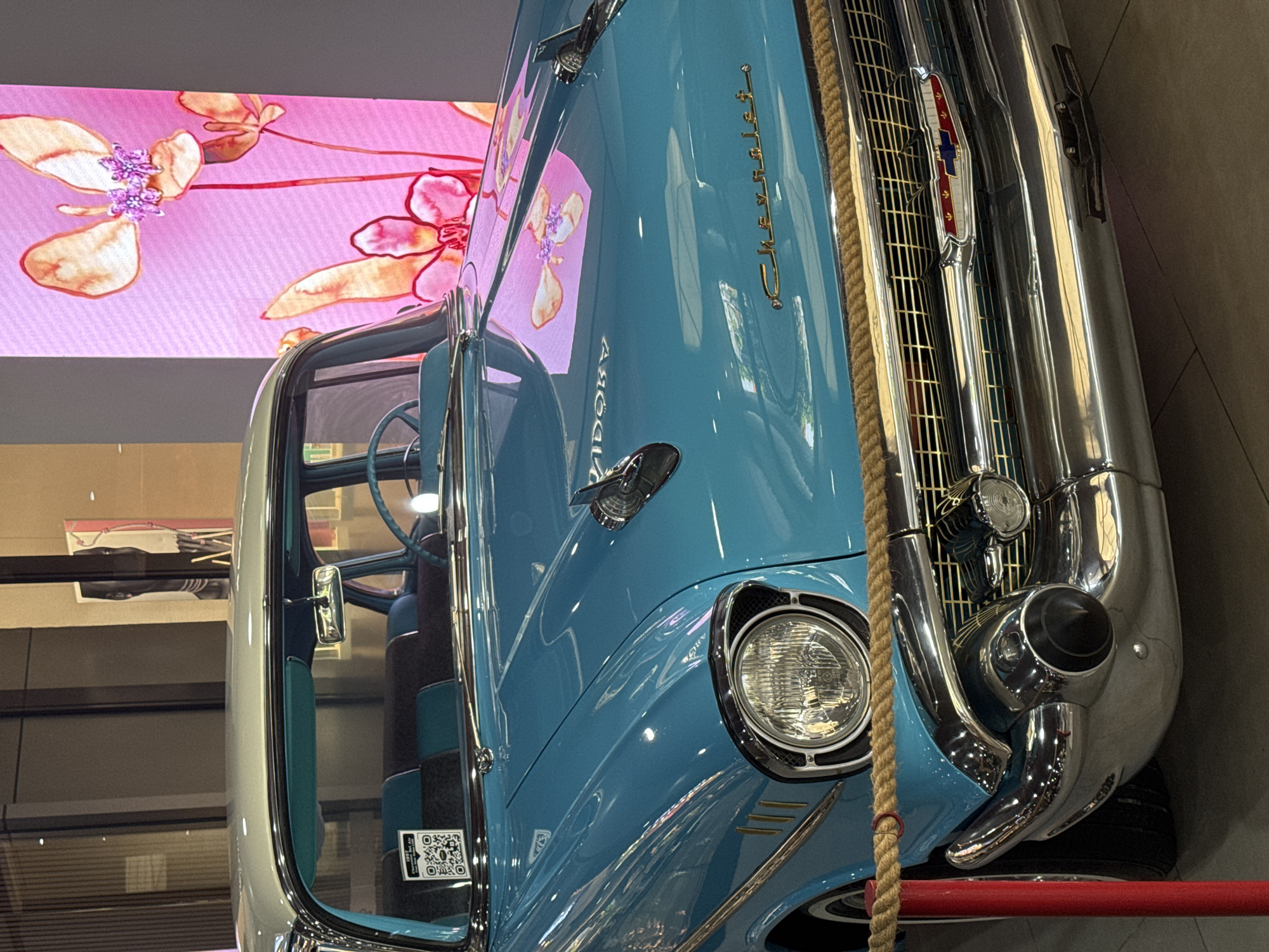 Here is a detailed description of the image:
The image depicts a close-up view of a vintage blue Chevrolet car, likely a 1957 Bel Air, from an unusual perspective. The car is partially obscured by a thick rope running vertically through the frame. The front of the car is prominent, showcasing the distinctive chrome grill, headlight, hood ornament, and Chevrolet script. The car's body is light blue.
Behind the car is a wall with a bright pink floral pattern. The interior of the car is visible, showing a two-tone blue and grey design on the seats and dashboard. There is a QR code sticker on the passenger-side window.
The overall environment suggests an indoor display or exhibit. The location information (city and state) is "Unknown, Unknown," as requested.