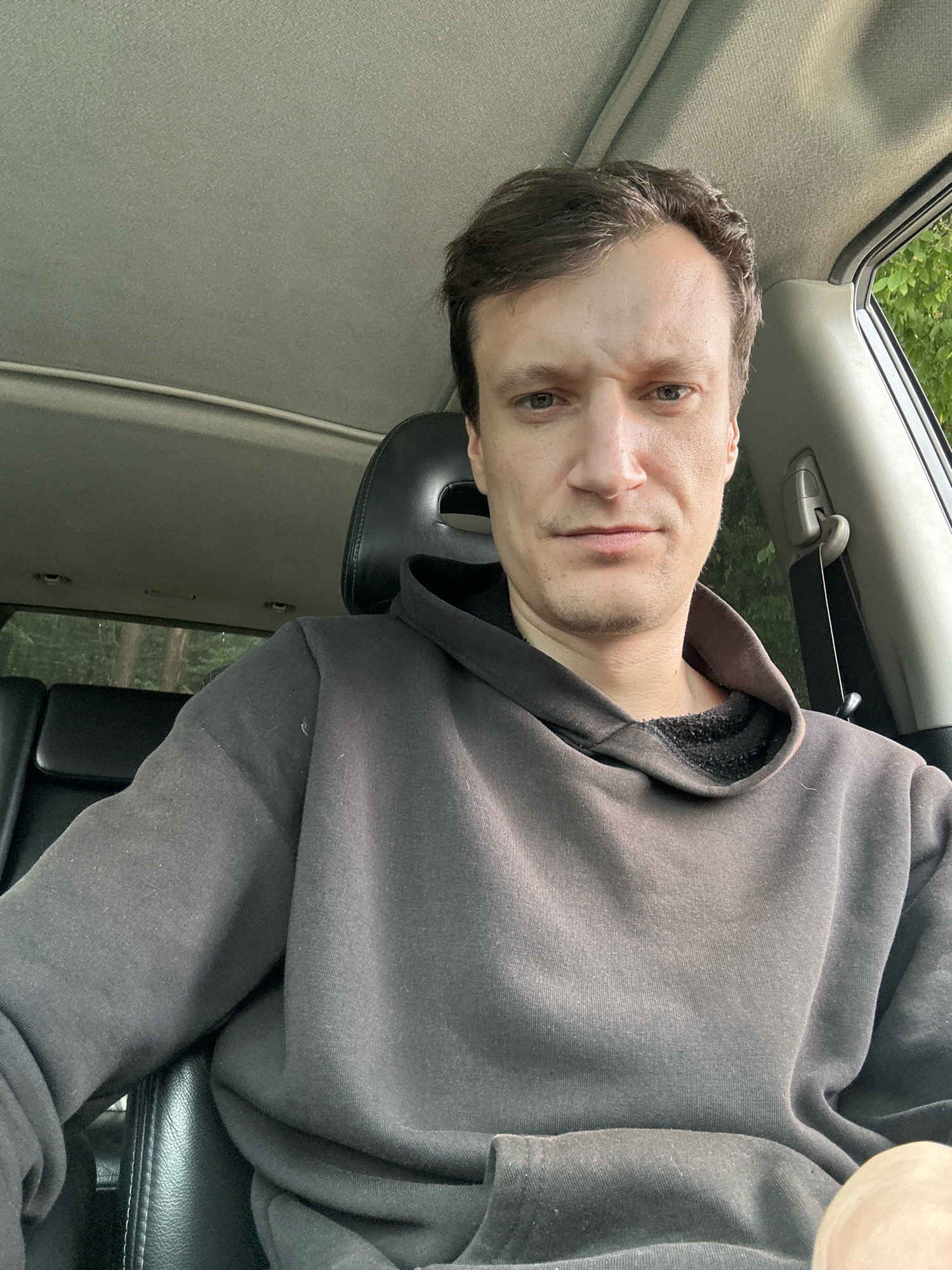 A man, appearing to be in his early 30s, is seated in the driver's seat of a vehicle, likely in Korchivka, Ukraine. He has short, dark brown hair and a fair complexion, with a slight stubble on his face. His expression is neutral, with his eyebrows slightly furrowed. He is wearing a dark gray hooded sweatshirt. The interior of the car is visible, including black leather seats, the car's headliner, and a portion of the window revealing greenery outside, suggesting a natural or wooded environment. The lighting indicates it is daytime.
