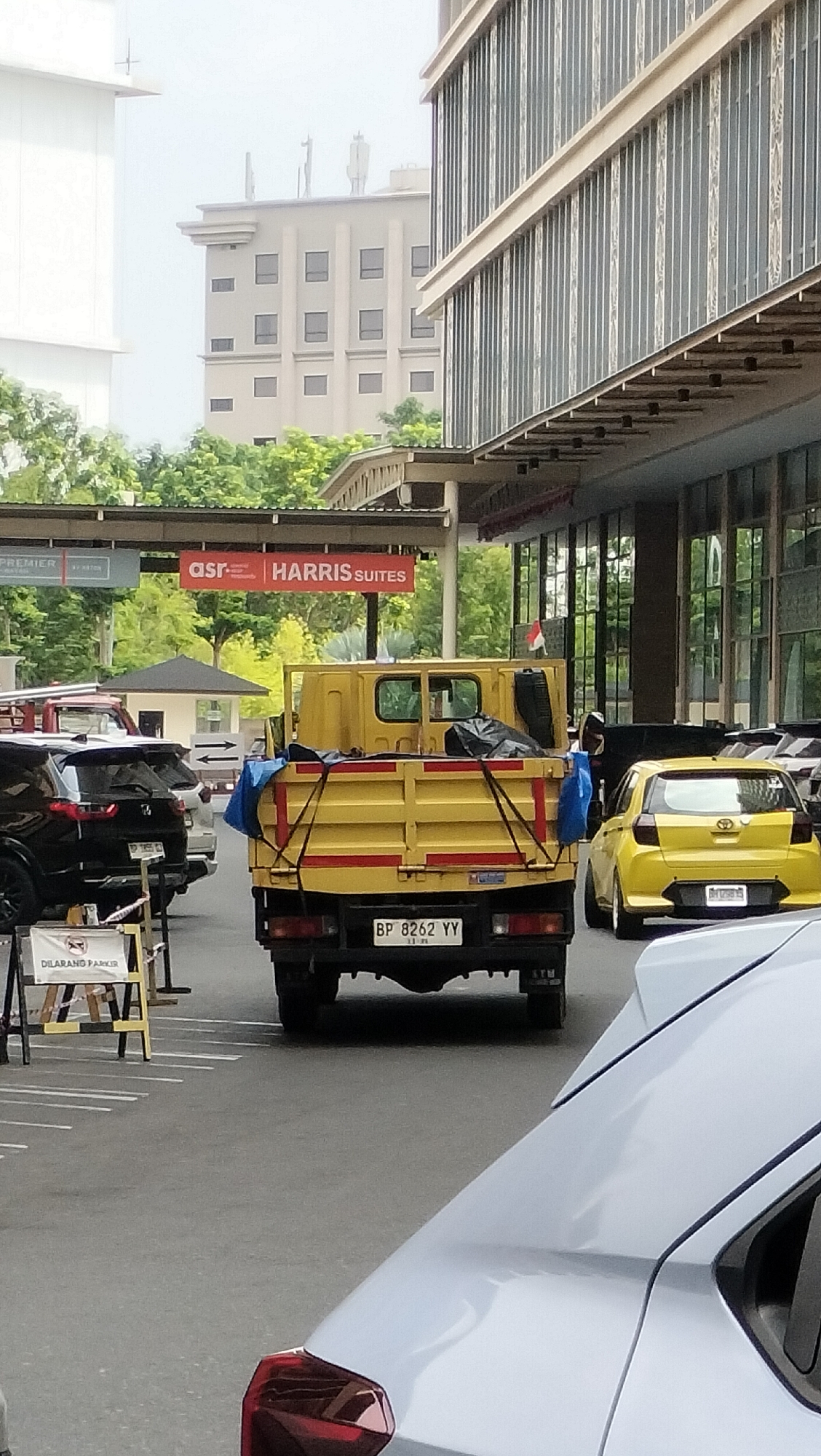 This is a photograph taken outdoors in Batam, Indonesia, likely in an area with commercial buildings and parking lots. In the foreground, the rear of a light gray car is visible, with its taillight and spoiler to the right and left, respectively. Behind this, a yellow flatbed truck is the central subject. It has a license plate that reads "BP 8262 YY". The truck bed is covered with a blue tarp and black plastic sheeting, obscuring its cargo. To the left of the truck, a black SUV is parked, and further to the left, a "DILARANG PARKIR" (No Parking) sign is visible. In the midground, a yellow hatchback car is parked to the right, with a license plate that is partially visible. Directly behind the truck, there is a small structure with a dark roof, possibly a guardhouse or kiosk. Above this, a red sign reads "asr HARRIS SUITES". In the background, several modern buildings with multiple windows are visible, along with lush green trees. The sky is overcast. The overall impression is an urban scene, possibly a hotel or commercial complex.