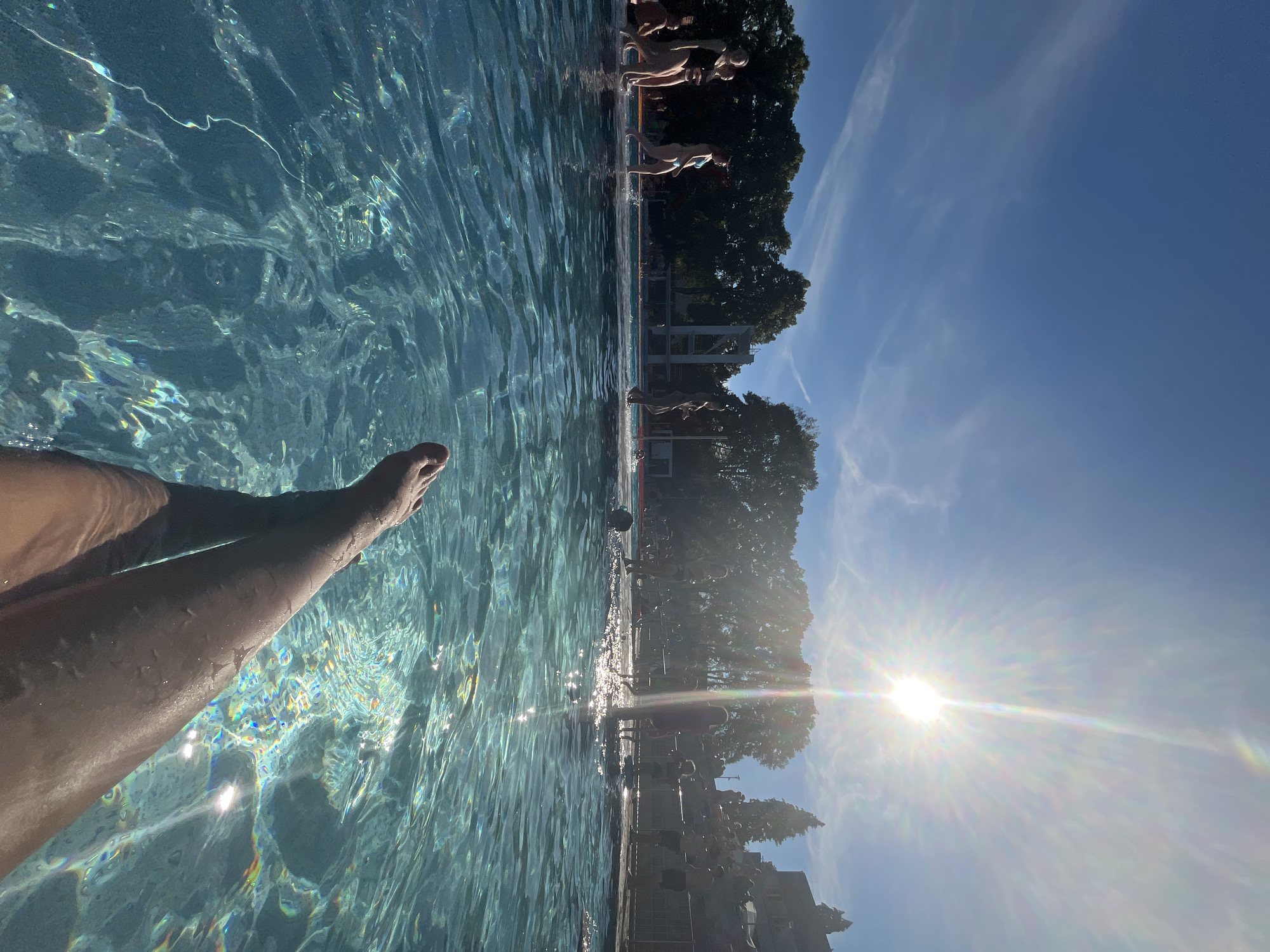 The image shows a first-person perspective of someone with their legs partially submerged in a swimming pool on a sunny day. The water is clear and reflects the sunlight, creating bright patterns. In the background, people are visible walking near the edge of the pool and in the shallow end. Trees and a building are seen beyond the pool area. The sky is blue with some wispy clouds. The sun is bright and high in the sky, causing a lens flare.